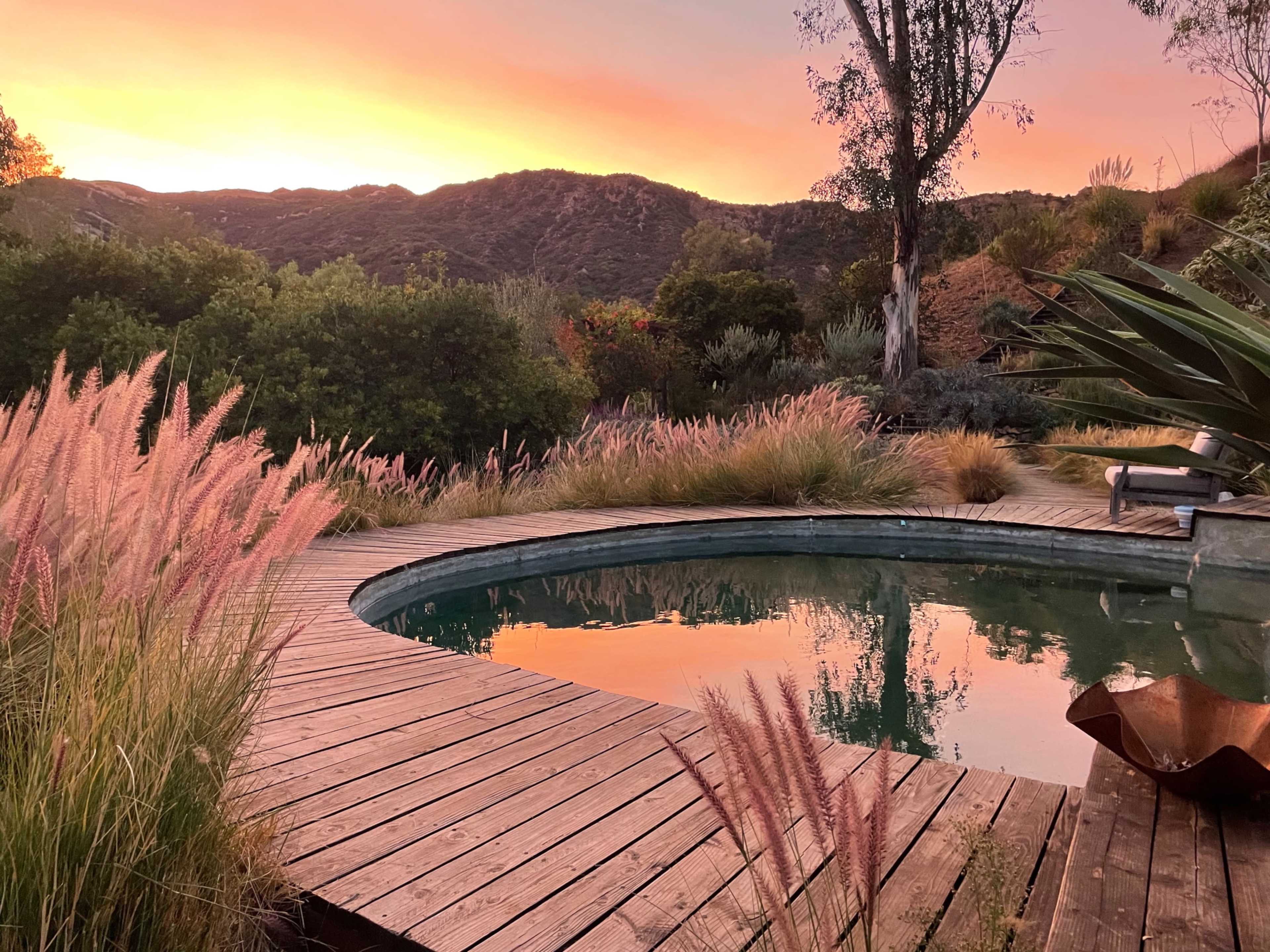 A swimming pool surrounded by wooden decking and tall grass reflects a colorful sunset backdrop over the hills.