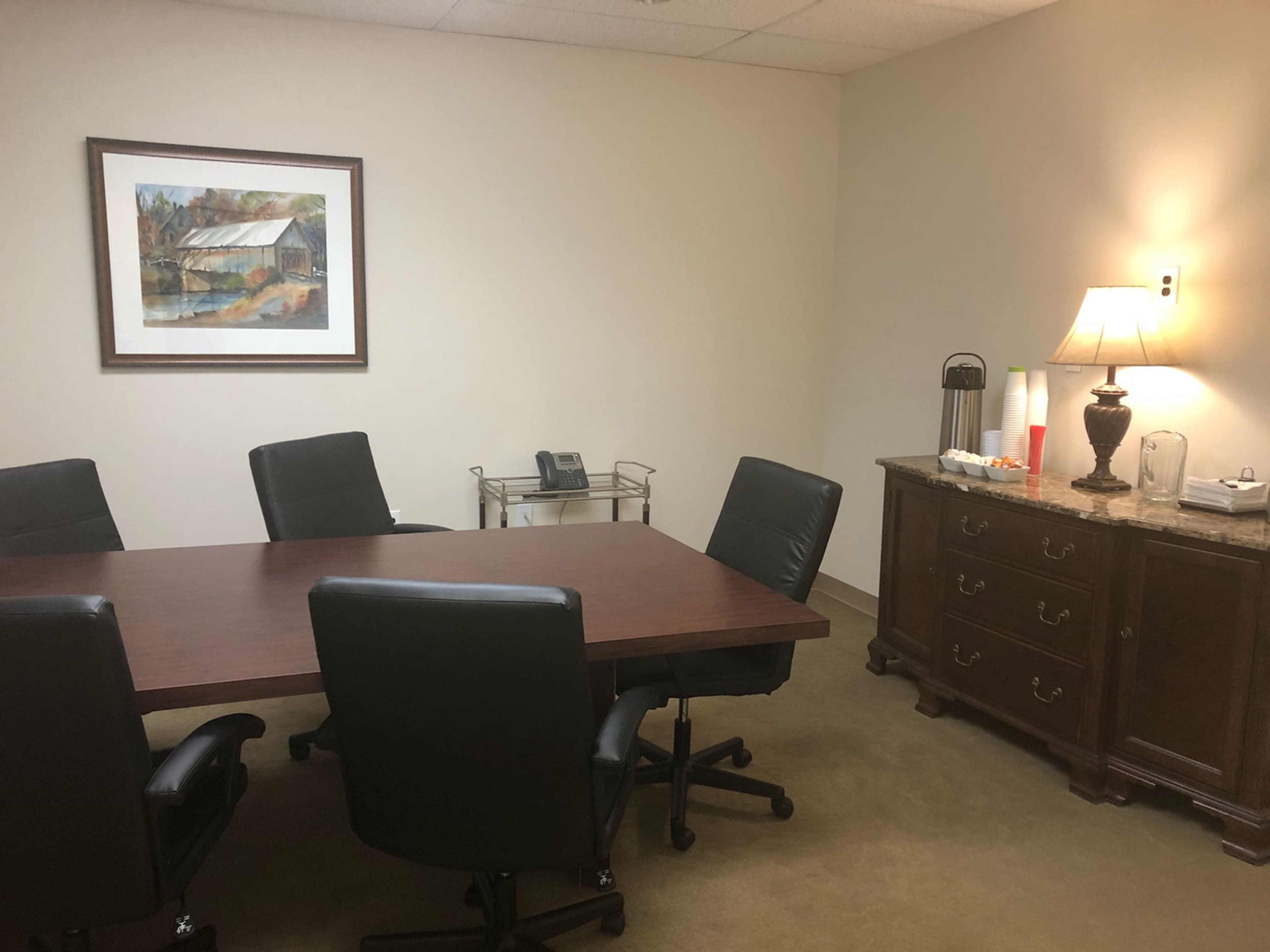 A small conference room with a wooden table surrounded by black chairs, a framed artwork on the wall, and a side table with a lamp and refreshments.