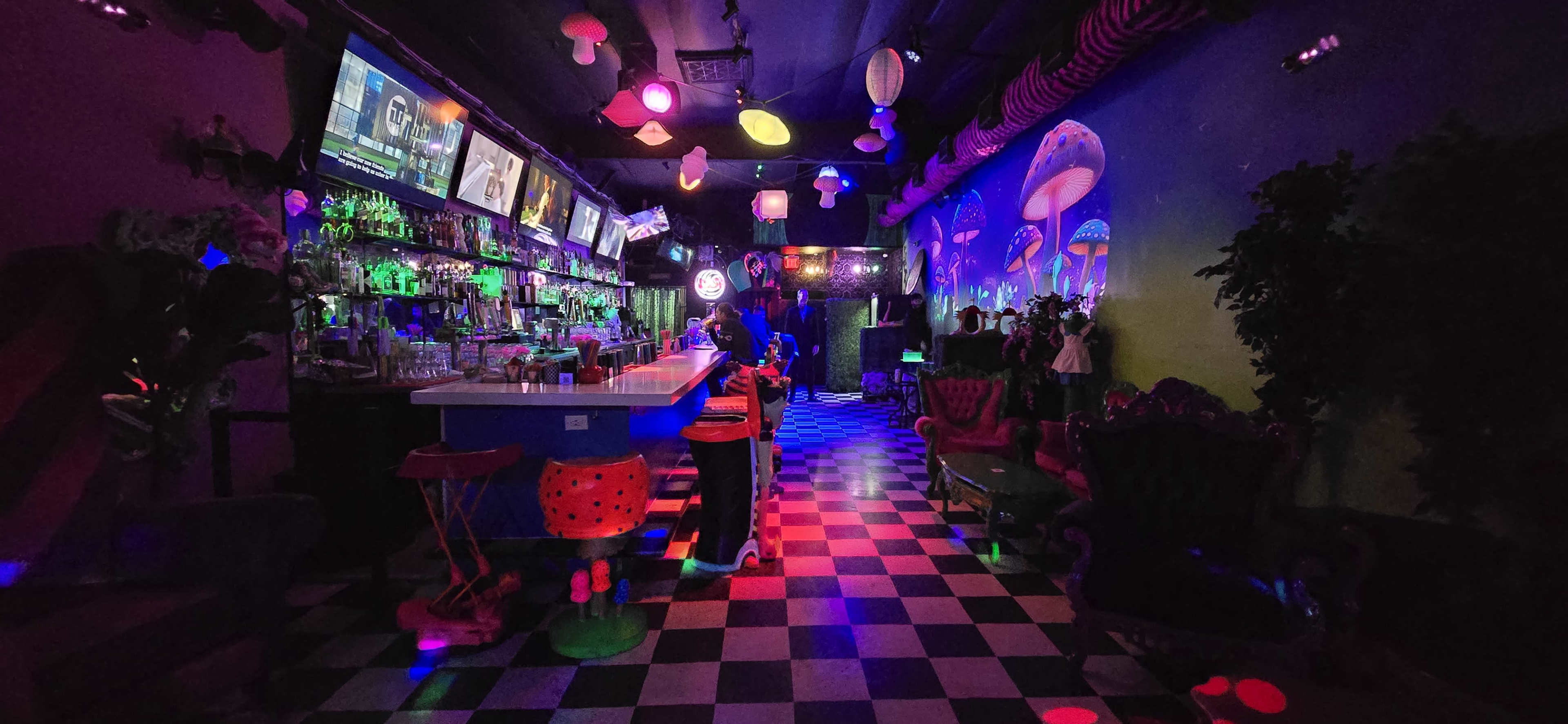 A dimly lit bar with a checkered floor, colorful lanterns, and multiple screens displaying content, featuring a long bar counter and eclectic seating arrangements.