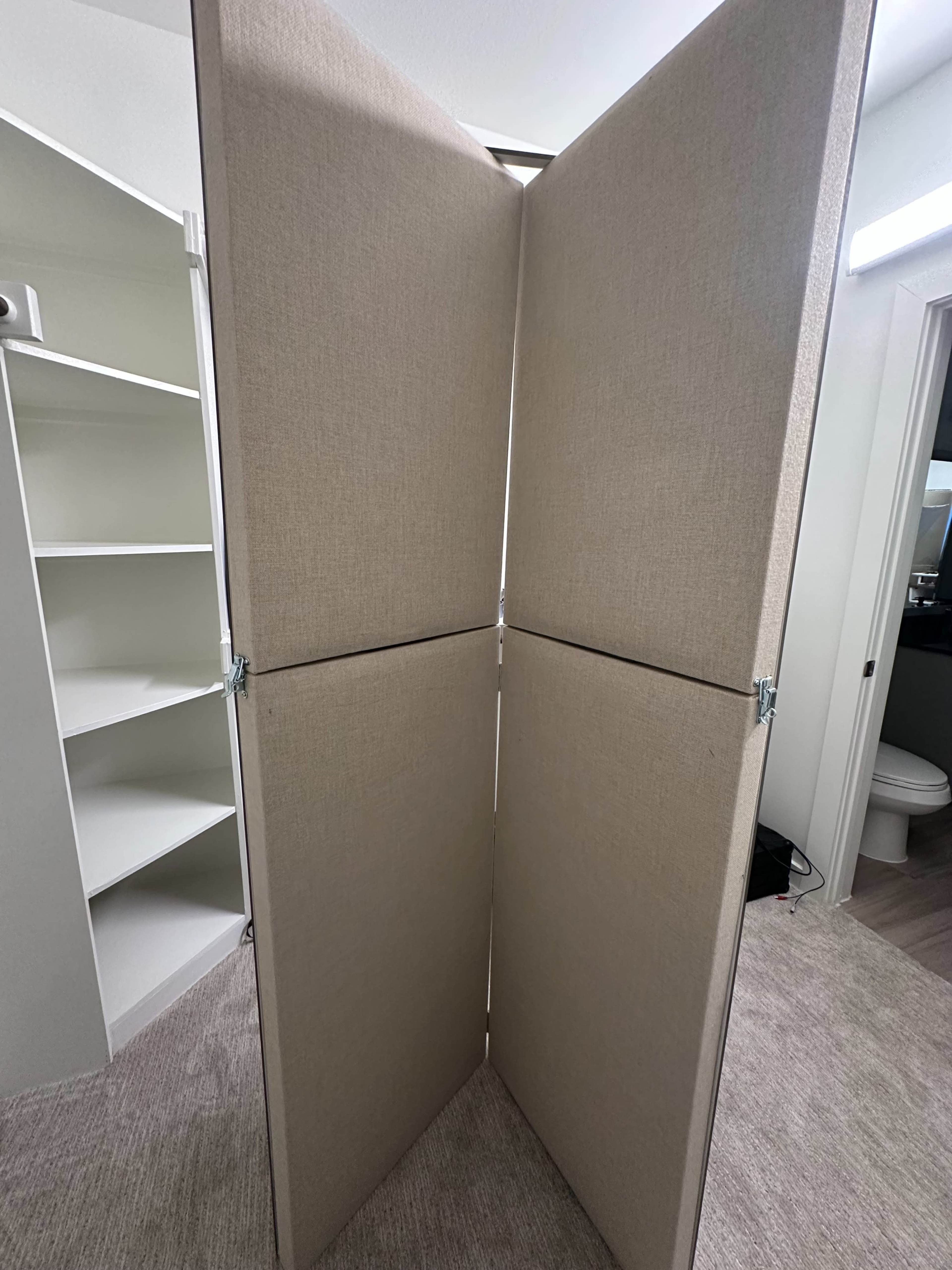 The image shows a folding room divider positioned in an empty space with a white shelving unit and a bathroom visible in the background.