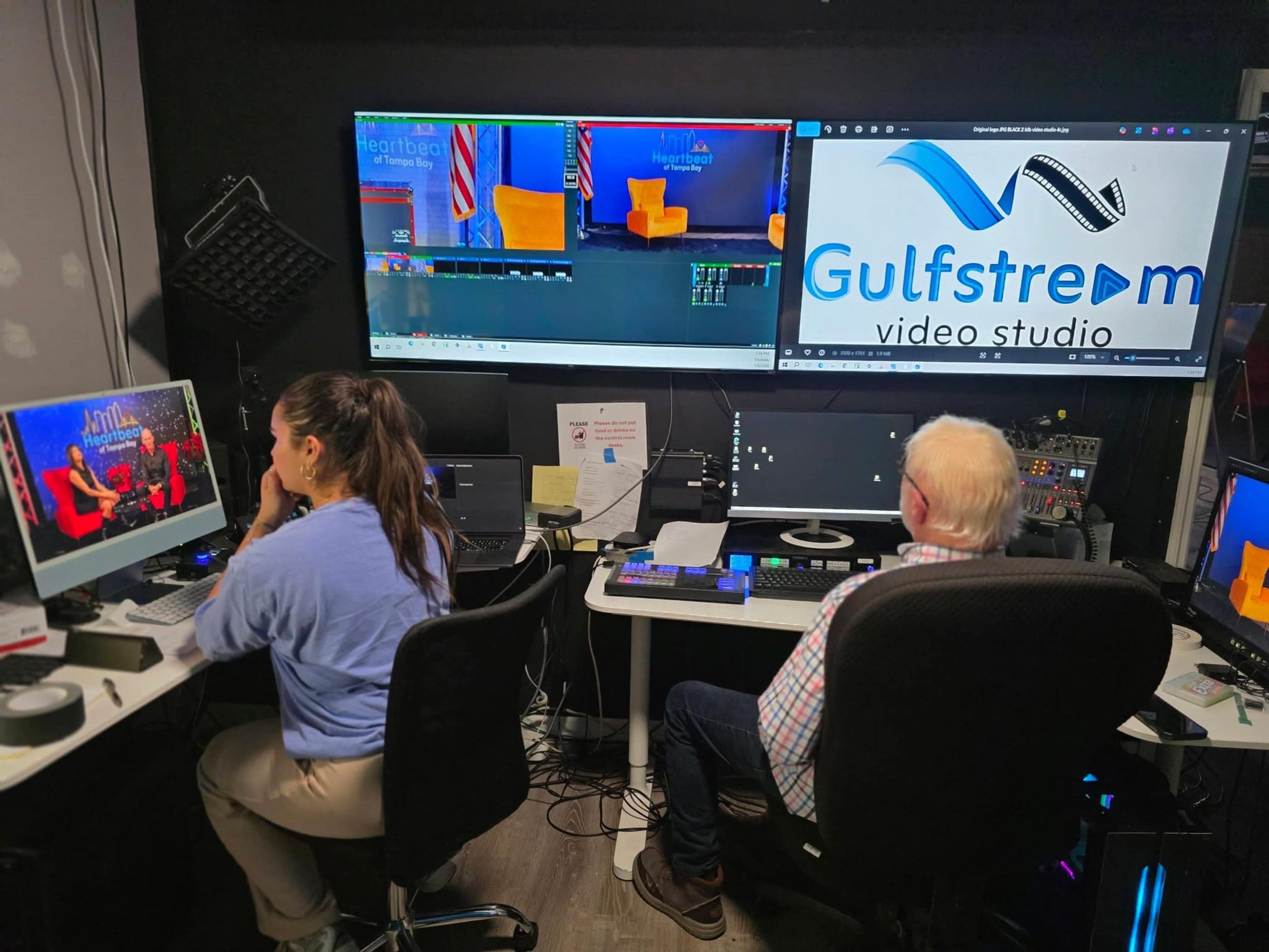 A woman operates a computer at a video editing workstation while a man monitors the output on large screens in a video studio.