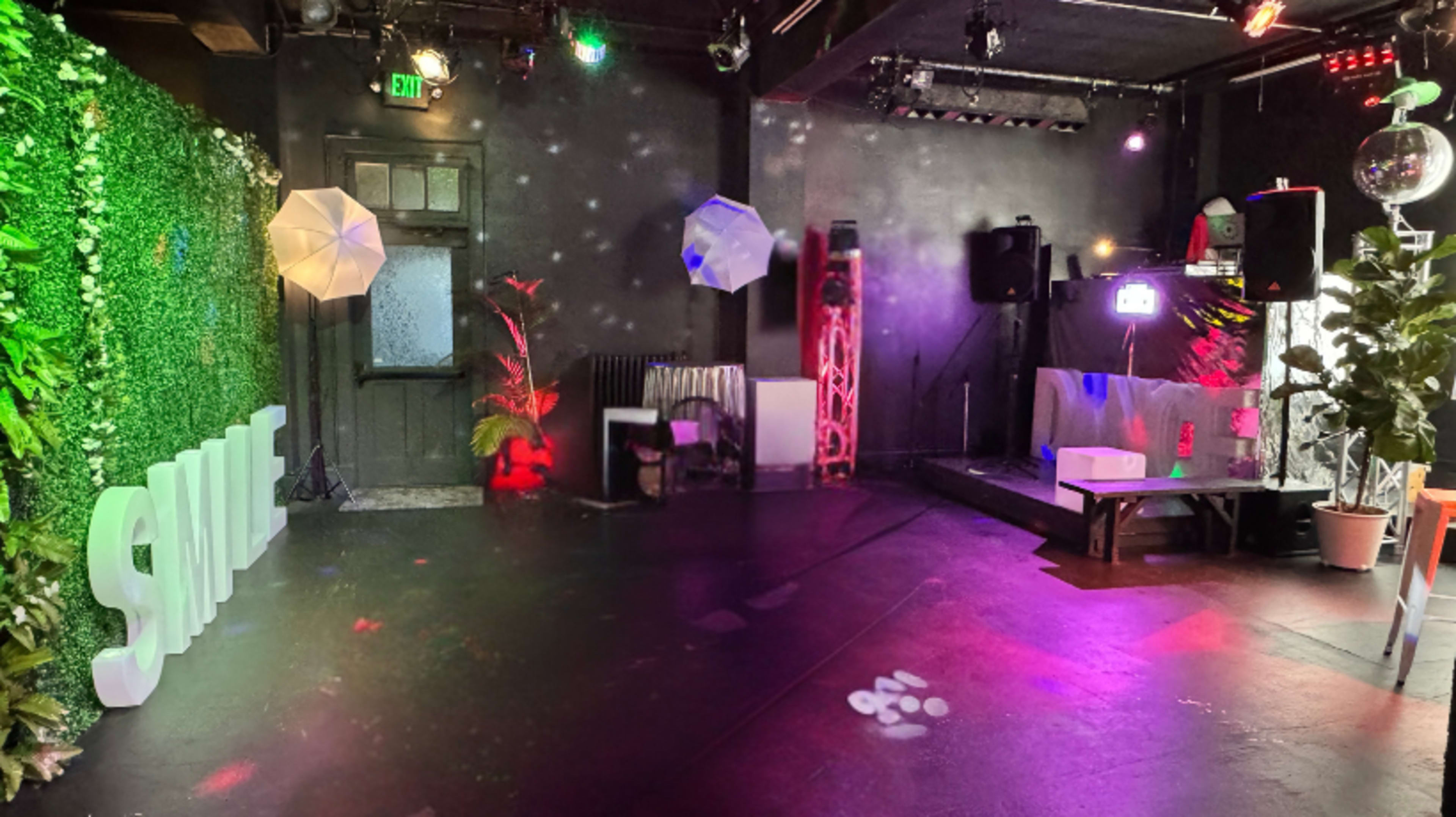 The image shows a dimly lit event space decorated with green foliage, illuminated umbrellas, and equipment for sound and lighting, featuring a backdrop that spells "SMILE."