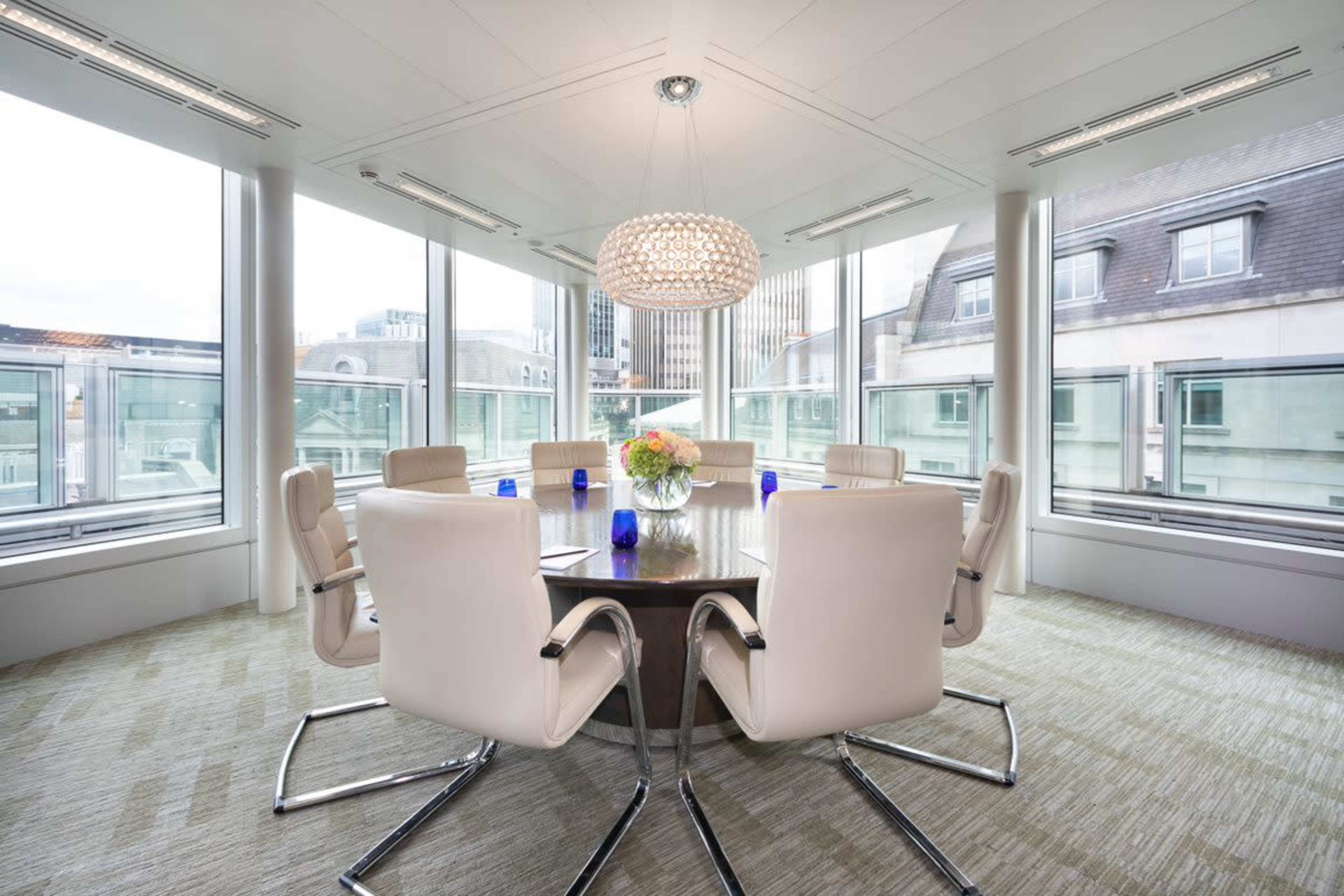 A conference room with a round table surrounded by white chairs and large windows offering a city view.