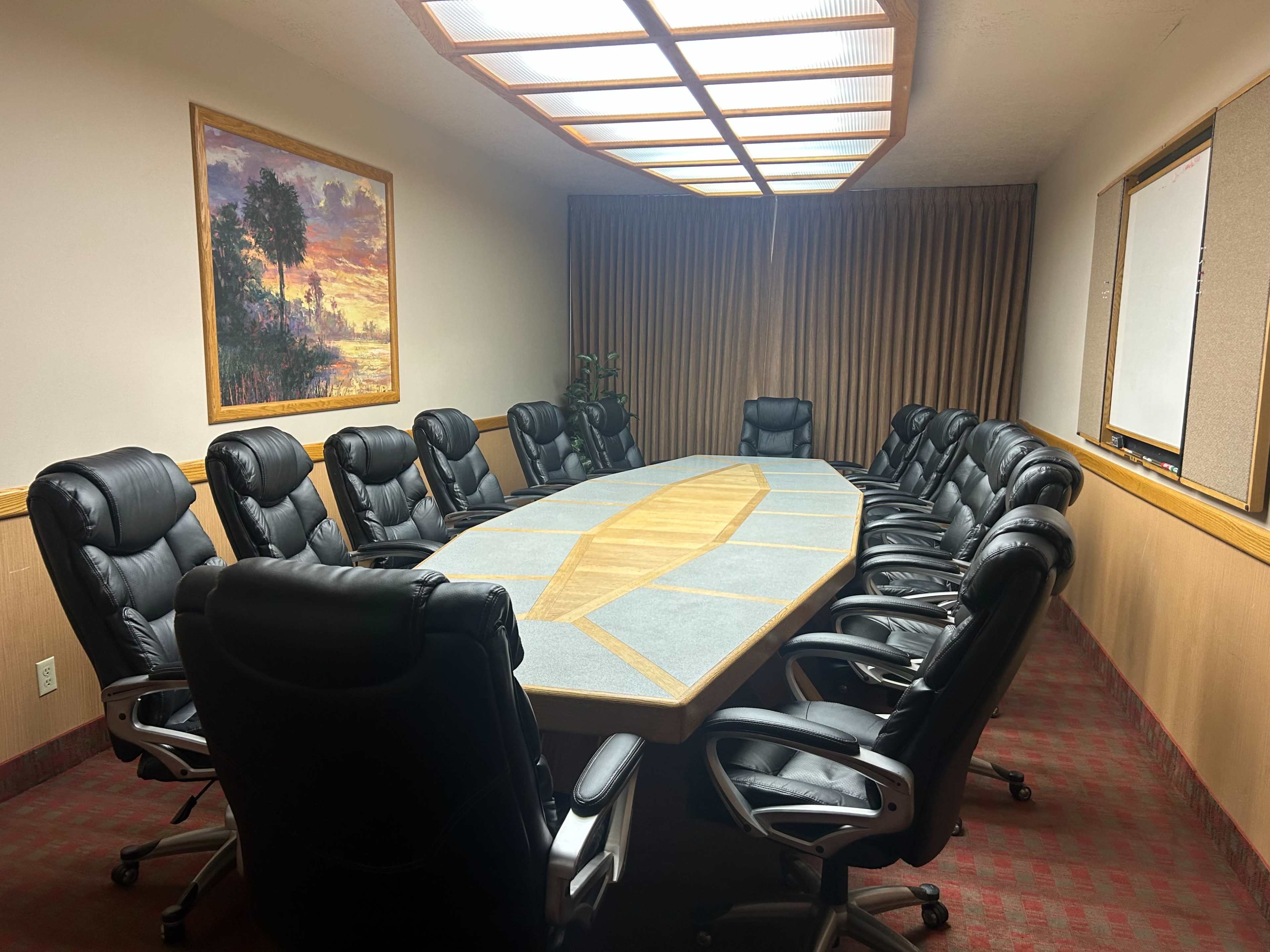 The image shows a conference room with a long oval table surrounded by sixteen black leather chairs and large windows above.