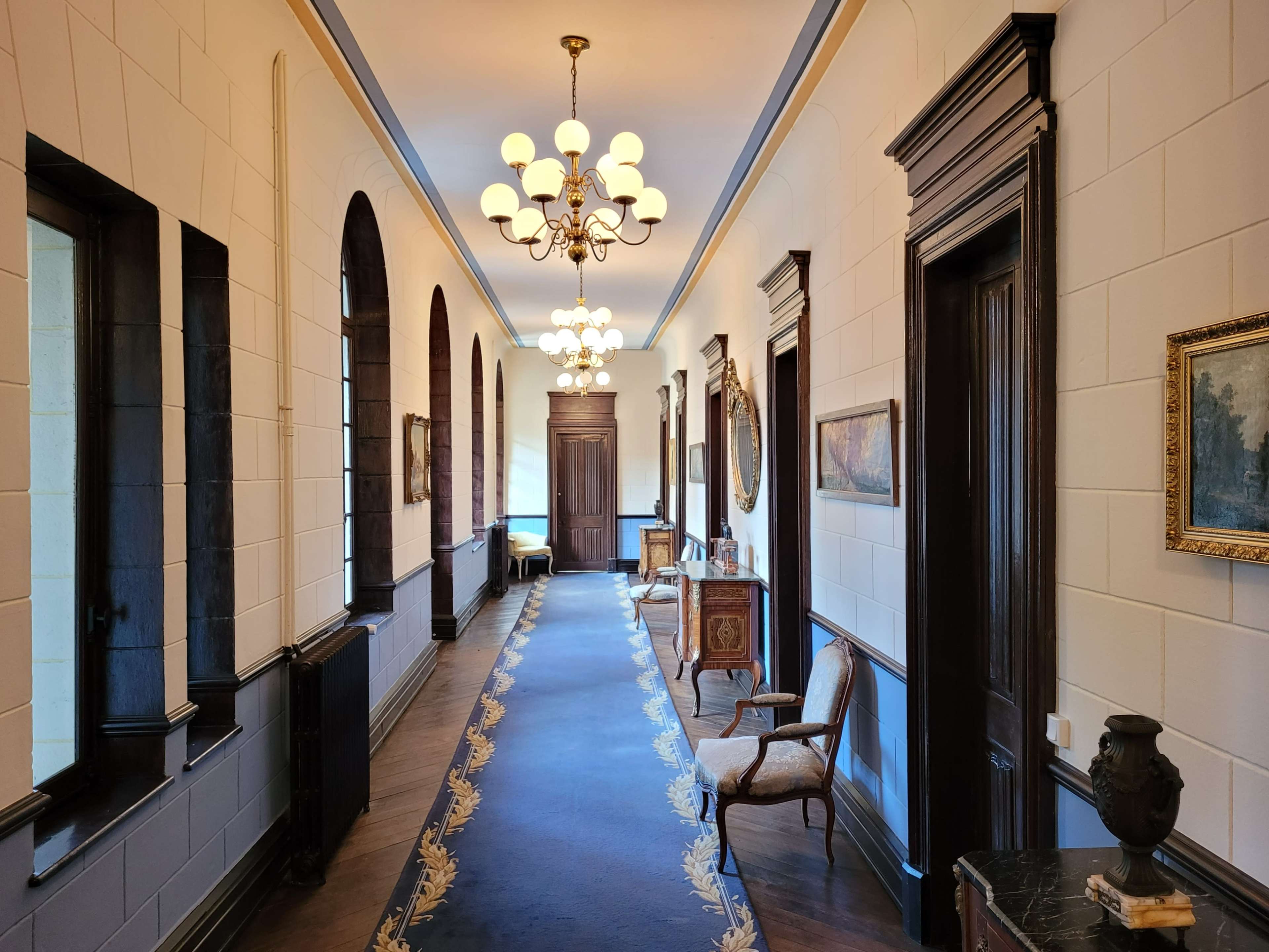 A long corridor adorned with arched windows, antique furniture, and hanging chandeliers, leading to a wooden door at the end.