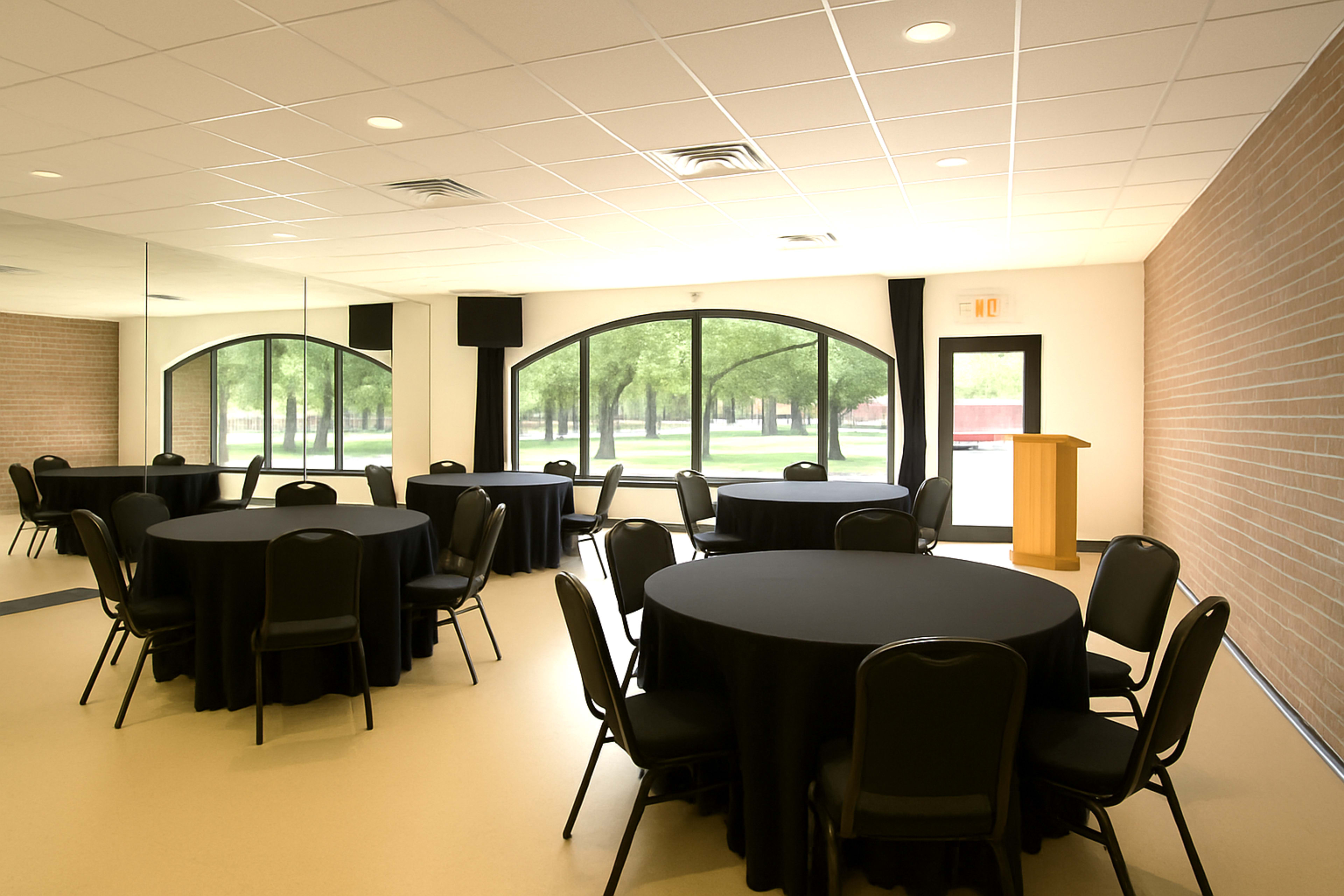 The image shows a spacious meeting room with round tables covered in black tablecloths, a wooden podium, and large windows overlooking a green outdoor area.
