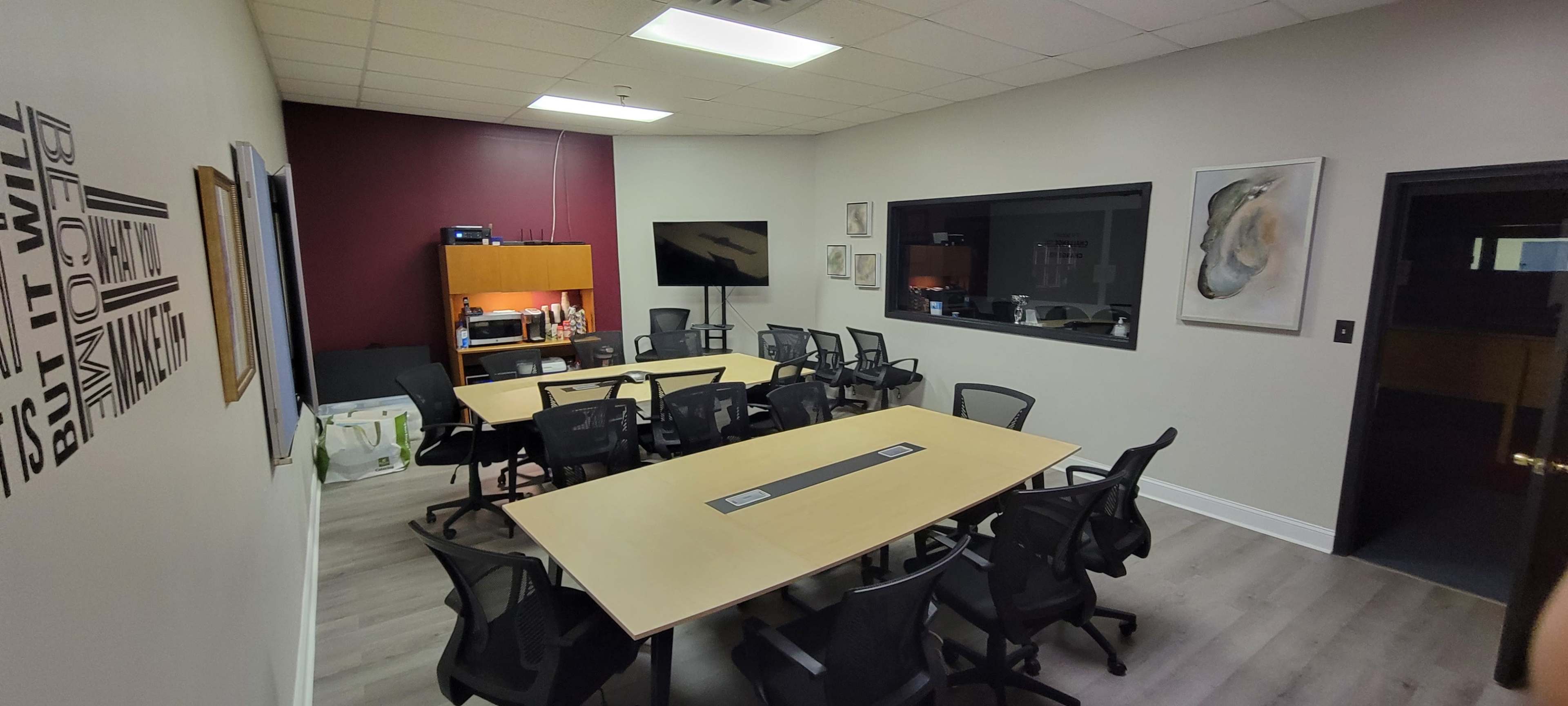 A conference room with a long table, multiple chairs, and a wall-mounted TV, with a large window displaying another room in the background.