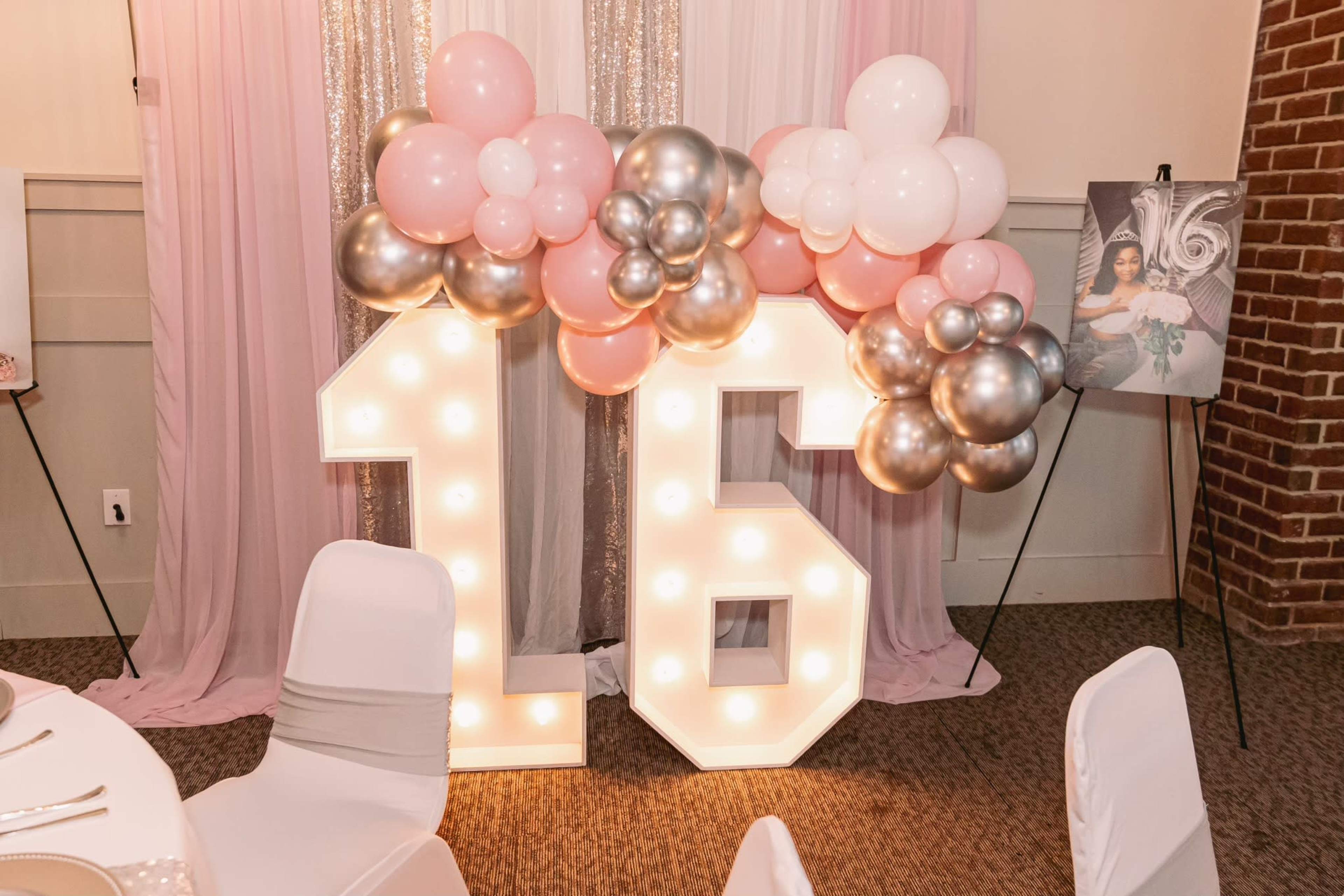 The image features large illuminated numbers "16" decorated with pink, white, and silver balloons, set against a backdrop of soft fabric draping.