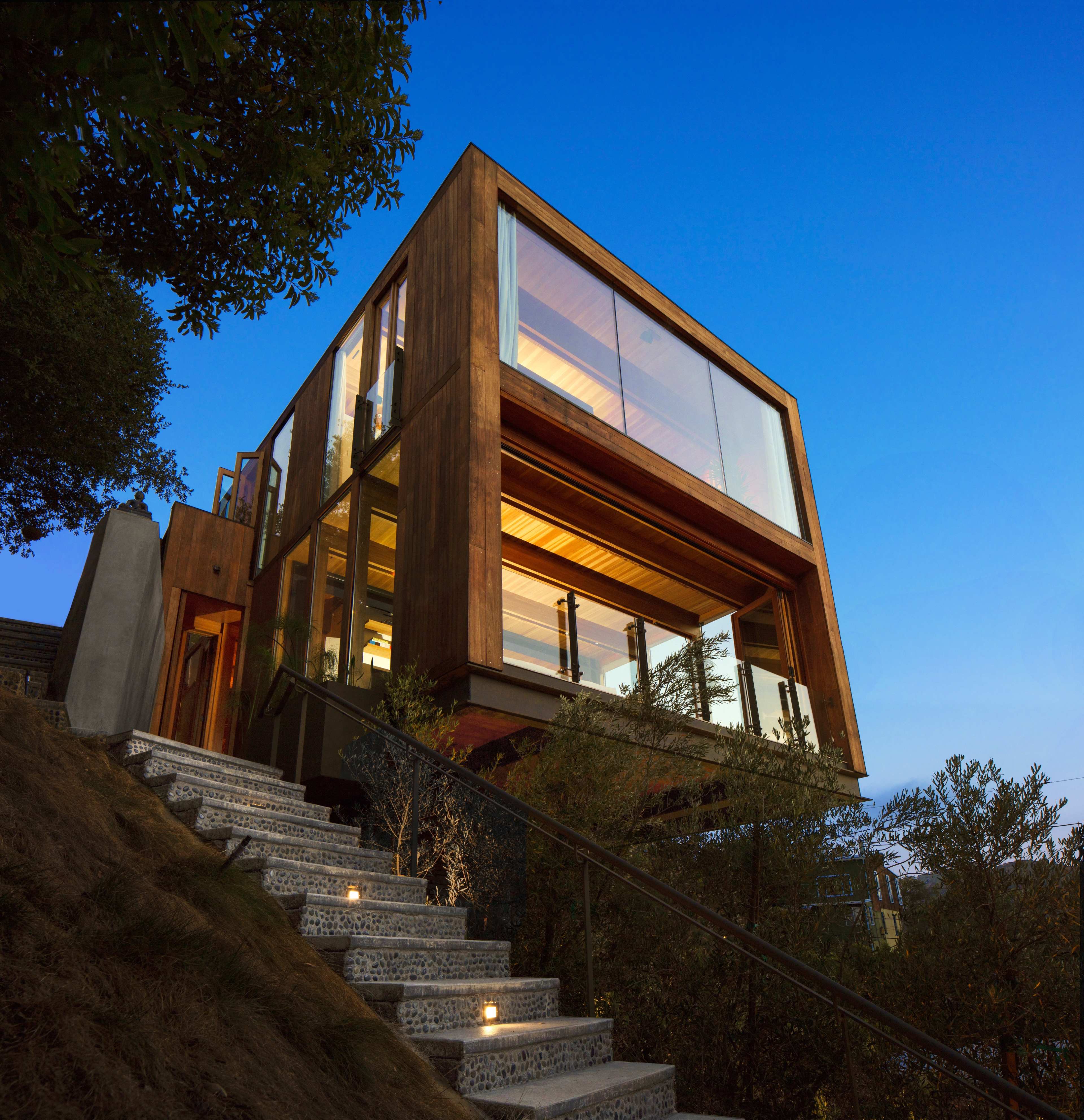 A modern wooden house with expansive glass windows is situated on a hillside, illuminated by outdoor lights along a stone pathway leading to its entrance.