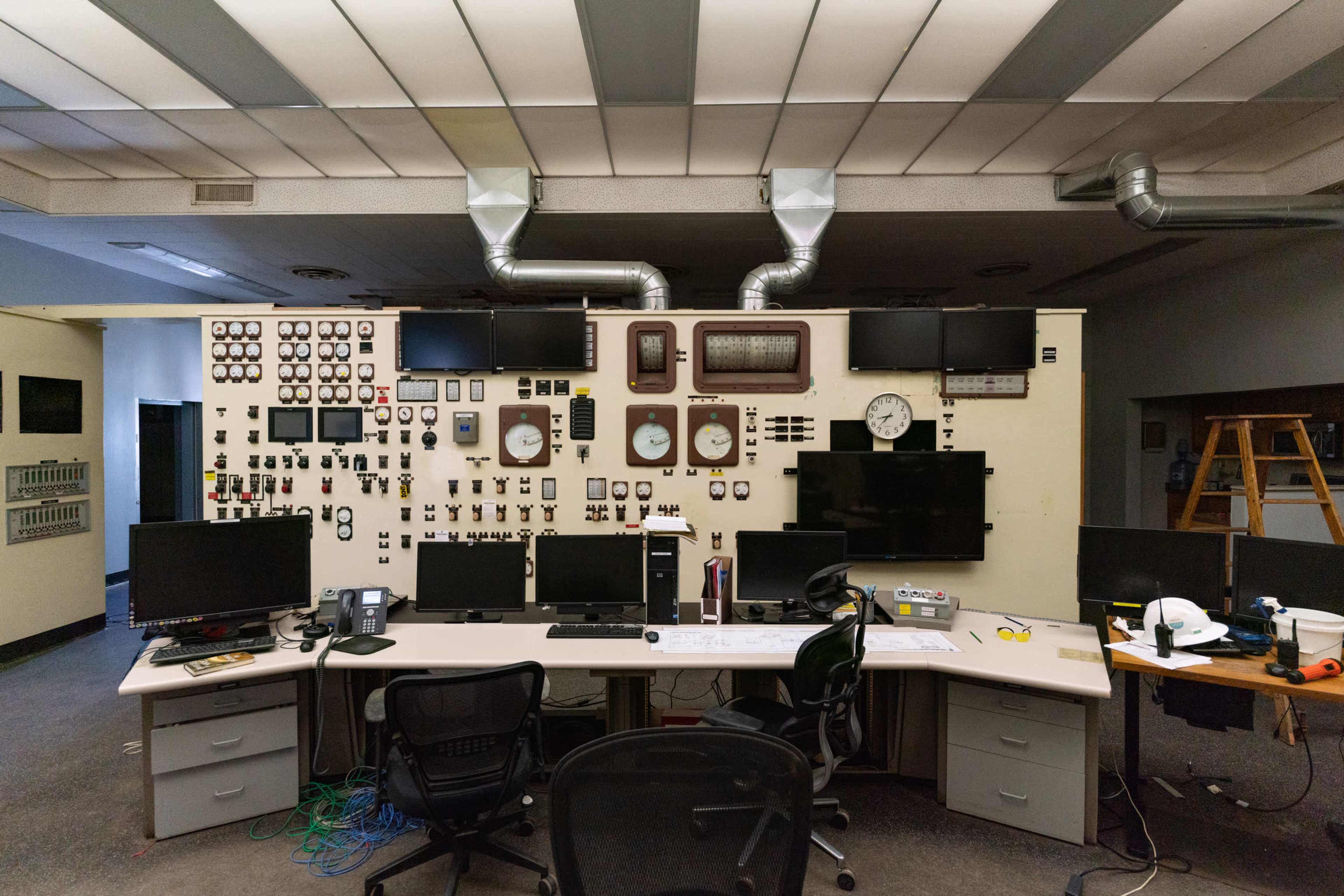 A control room with a large wall displaying various gauges, clocks, and monitors, along with a central desk equipped with multiple computer screens and chairs.