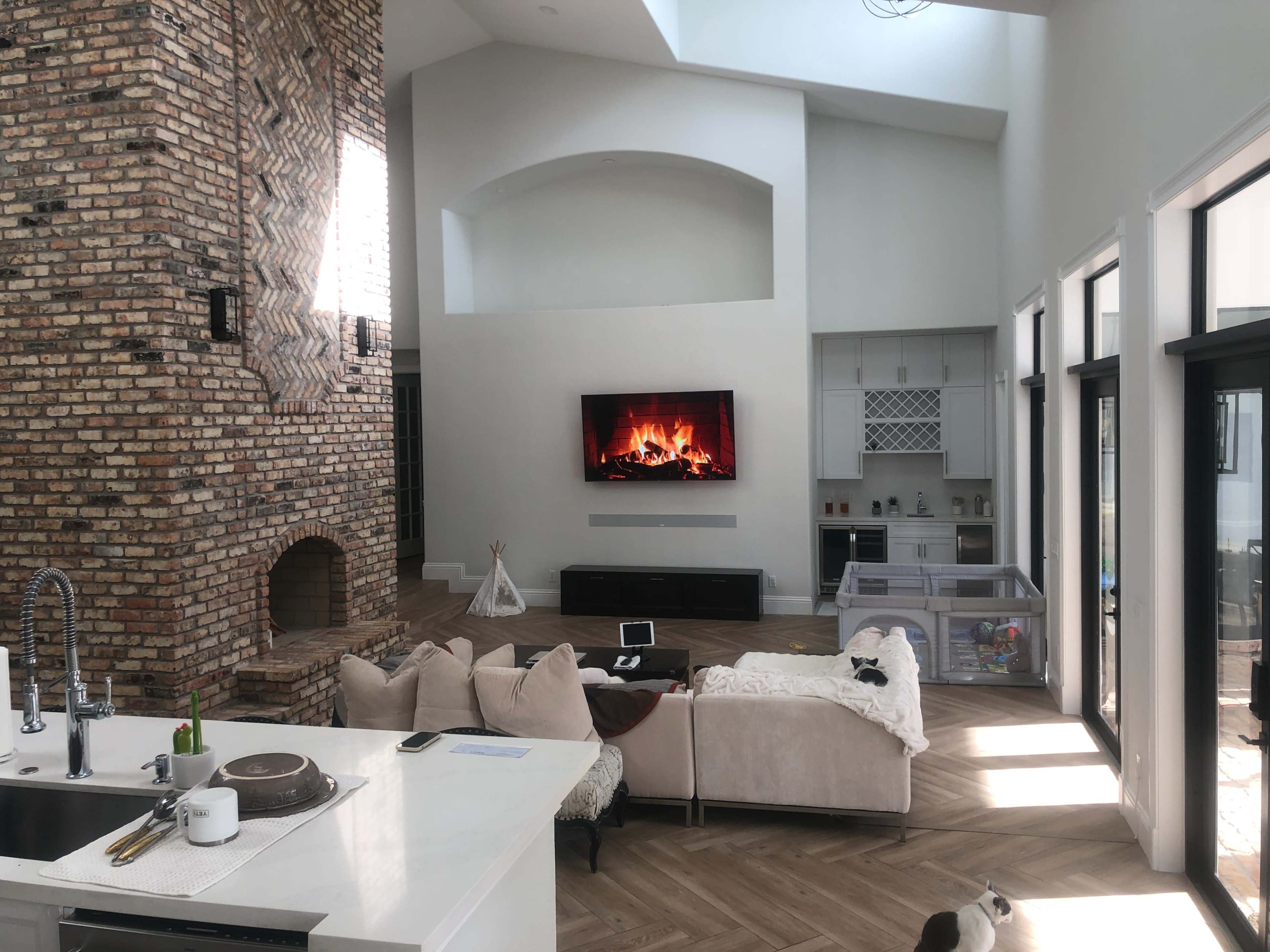 A spacious living room features a tall stone fireplace, a large TV mounted on the wall, and a light-colored sofa facing a wood floor.