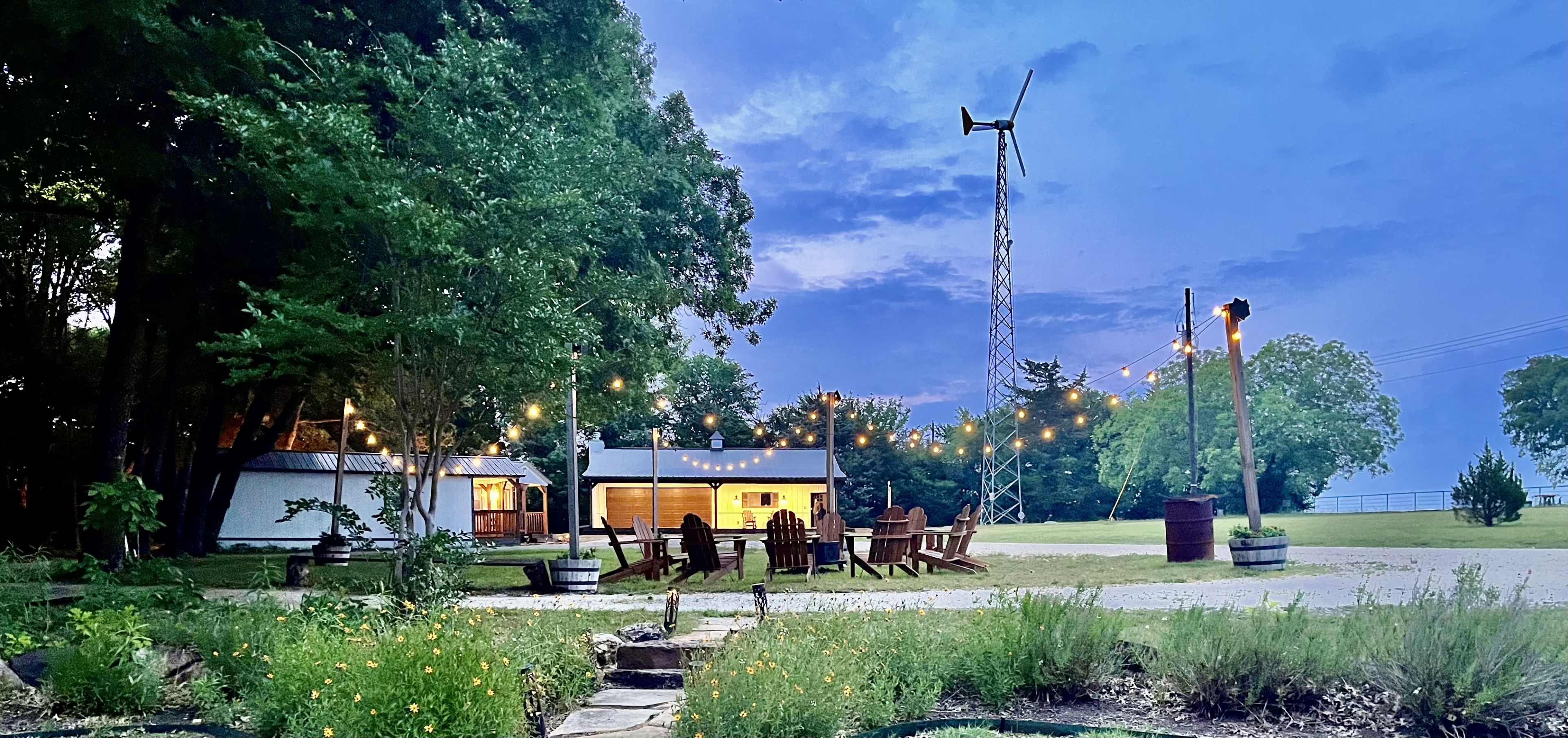 A modern outdoor space features string lights, wooden chairs arranged around a fire pit, and a wind turbine in the background at dusk.