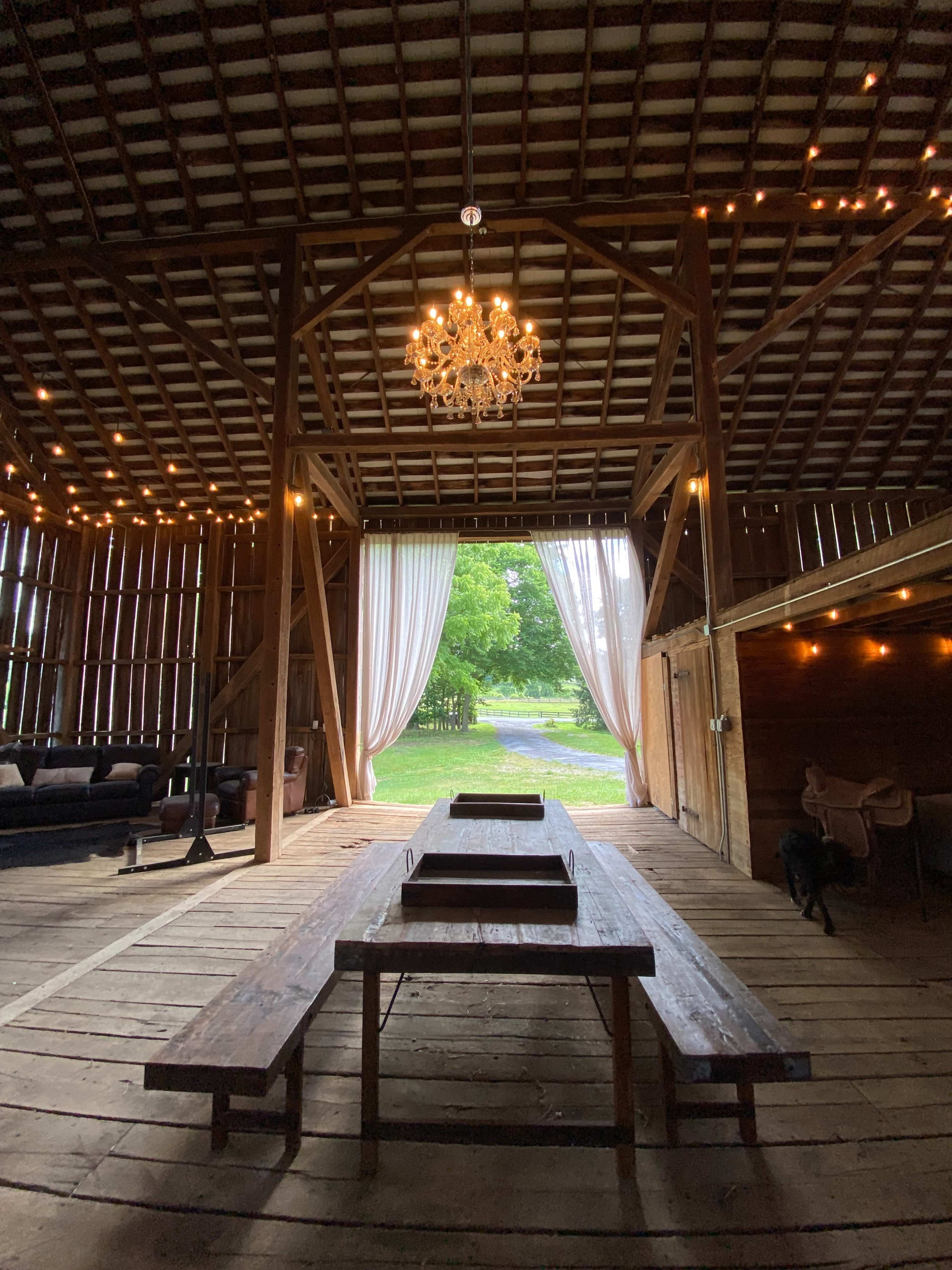 The interior of a spacious barn with wooden flooring, a long table in the center, and large windows draped with sheer curtains that open to a green outdoor area.