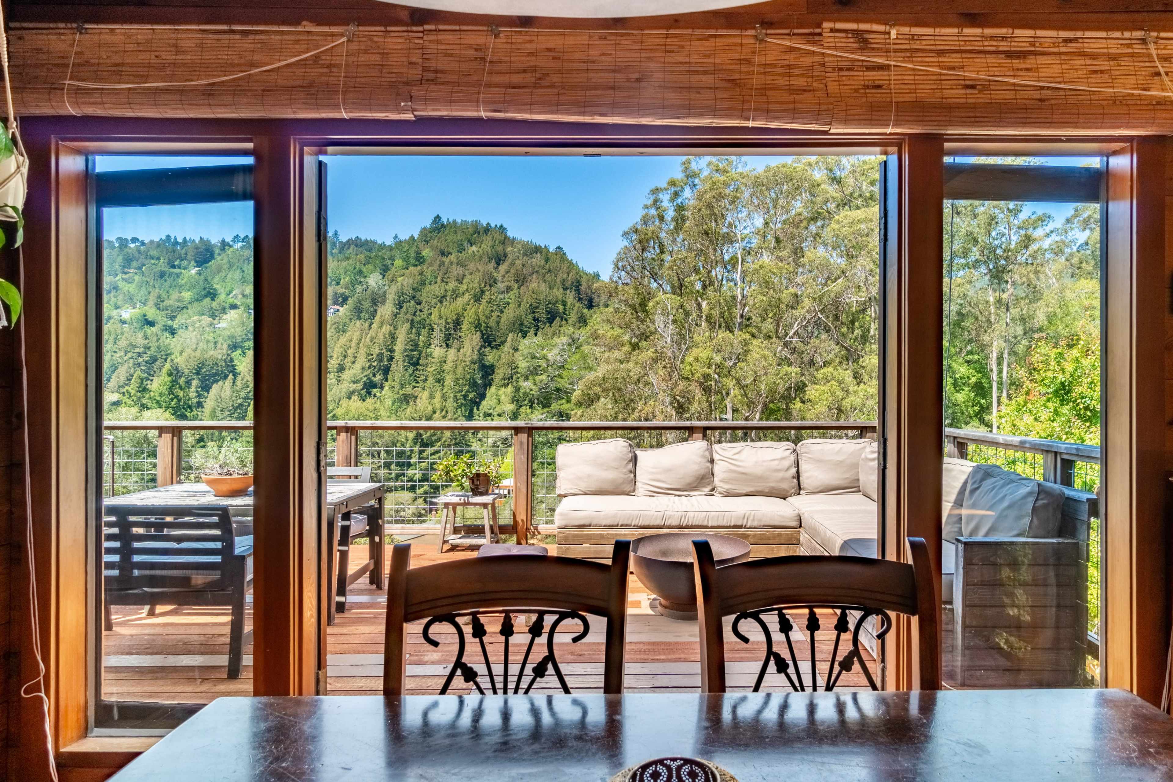 A living space with large windows overlooking a deck that features outdoor seating and a table, surrounded by trees and hills.
