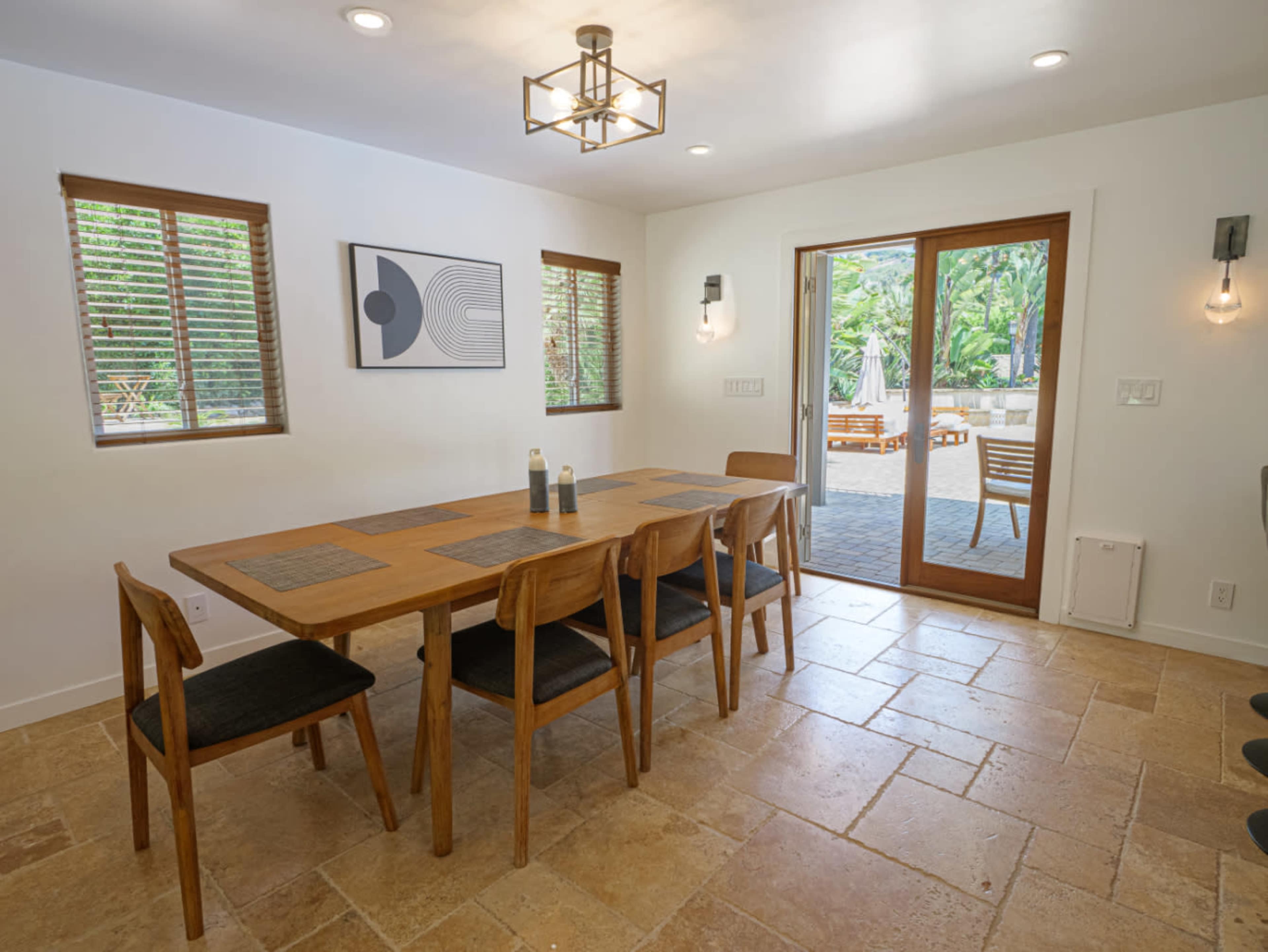A dining room features a long wooden table surrounded by chairs, with large sliding doors that open to a patio area and windows allowing natural light.