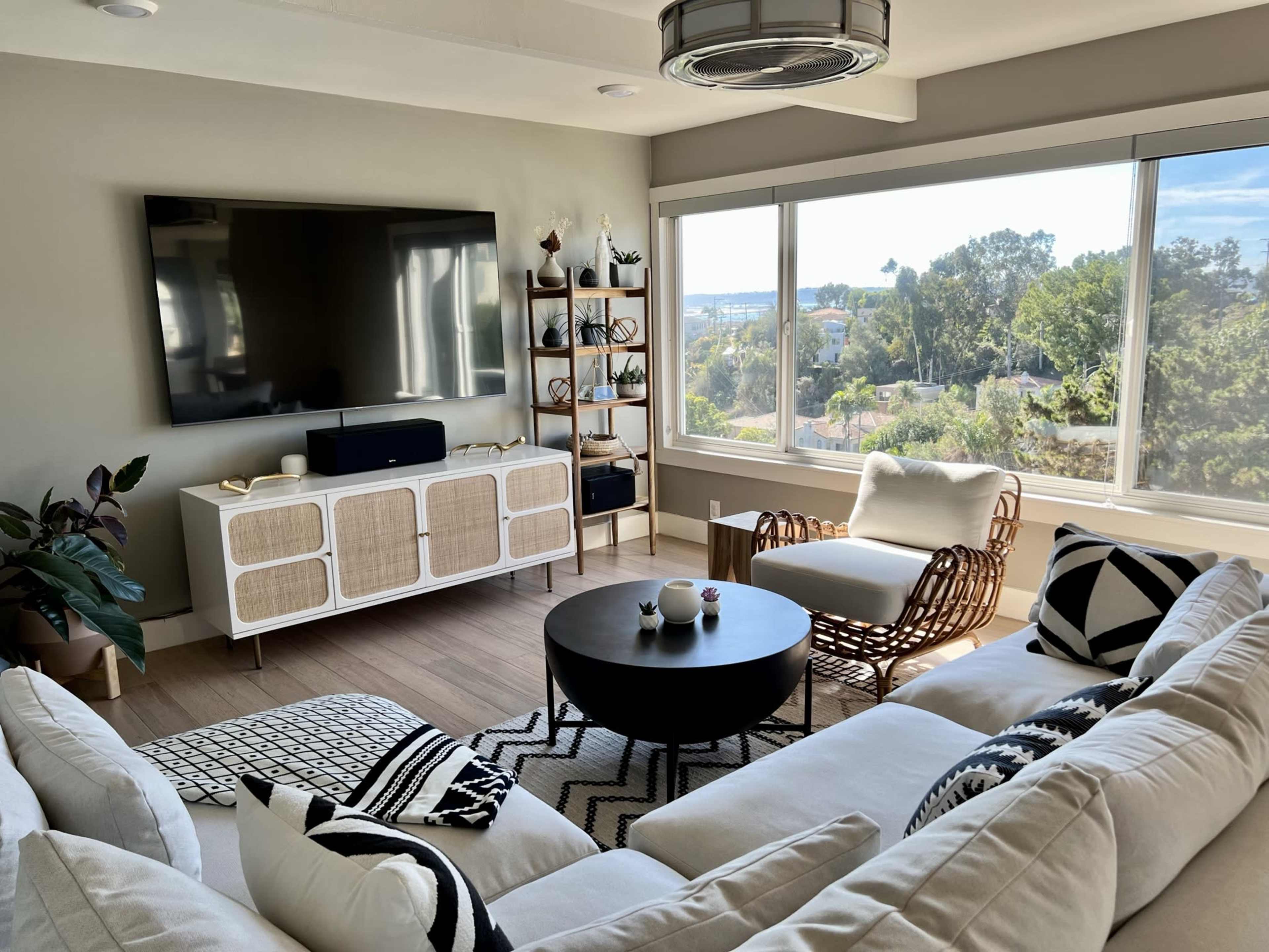 The image shows a modern living room with a large window, a television on the wall, a bookshelf, and a white sectional sofa arranged around a circular coffee table.
