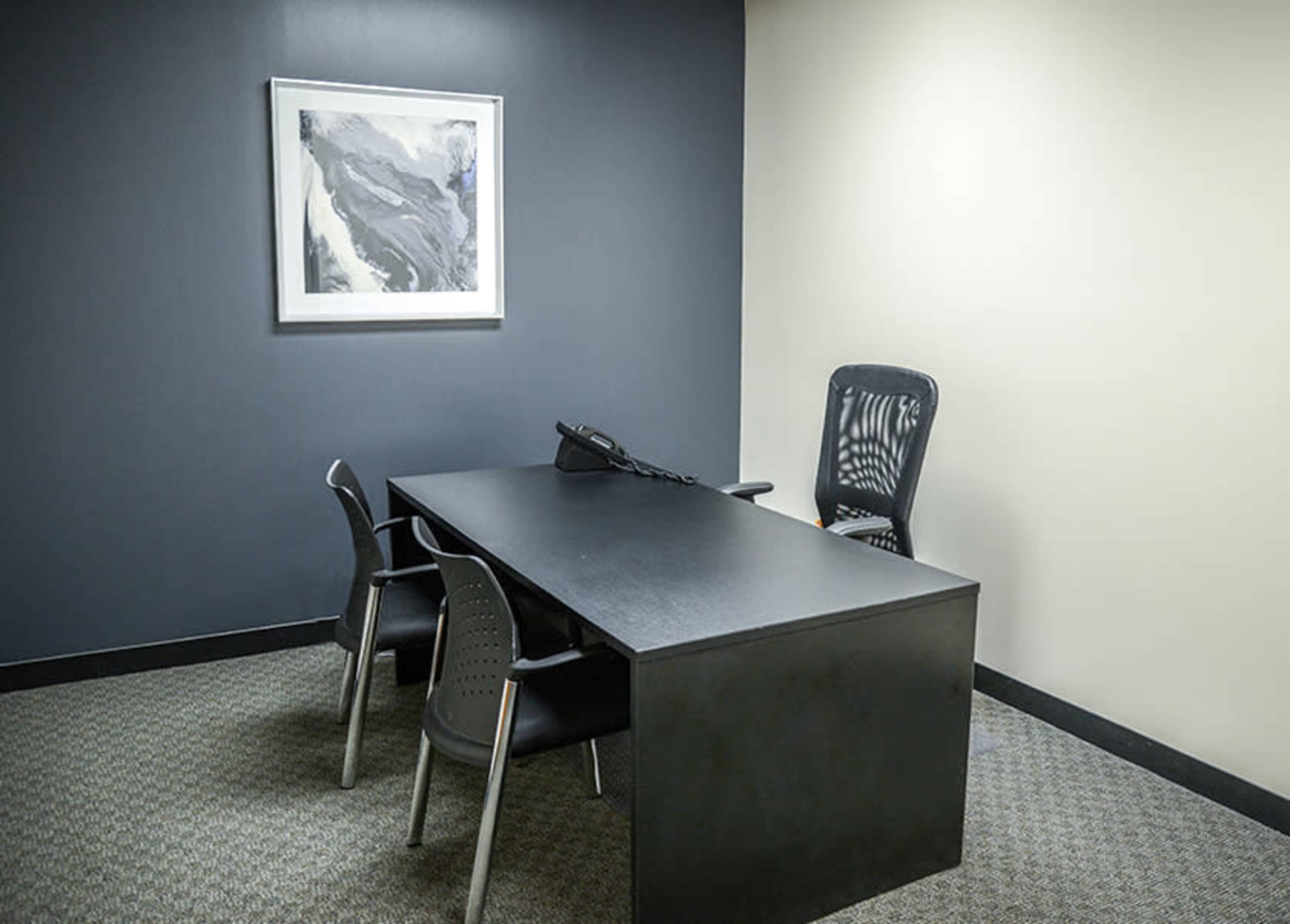A small office space with a black desk, three chairs, a telephone, and a piece of artwork on the wall.