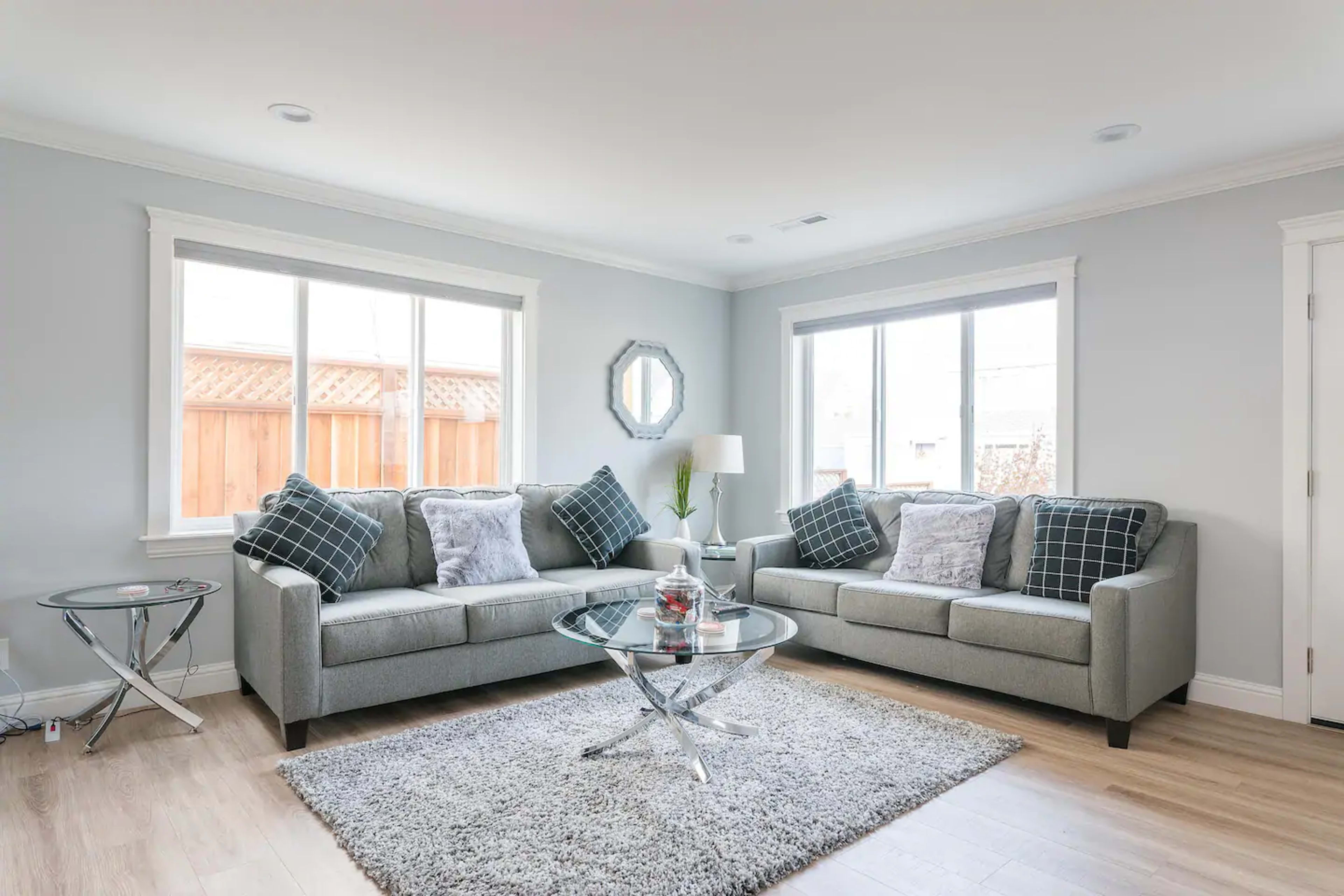 A modern living room features two gray sofas with plaid pillows, a circular glass coffee table, and large windows allowing natural light.