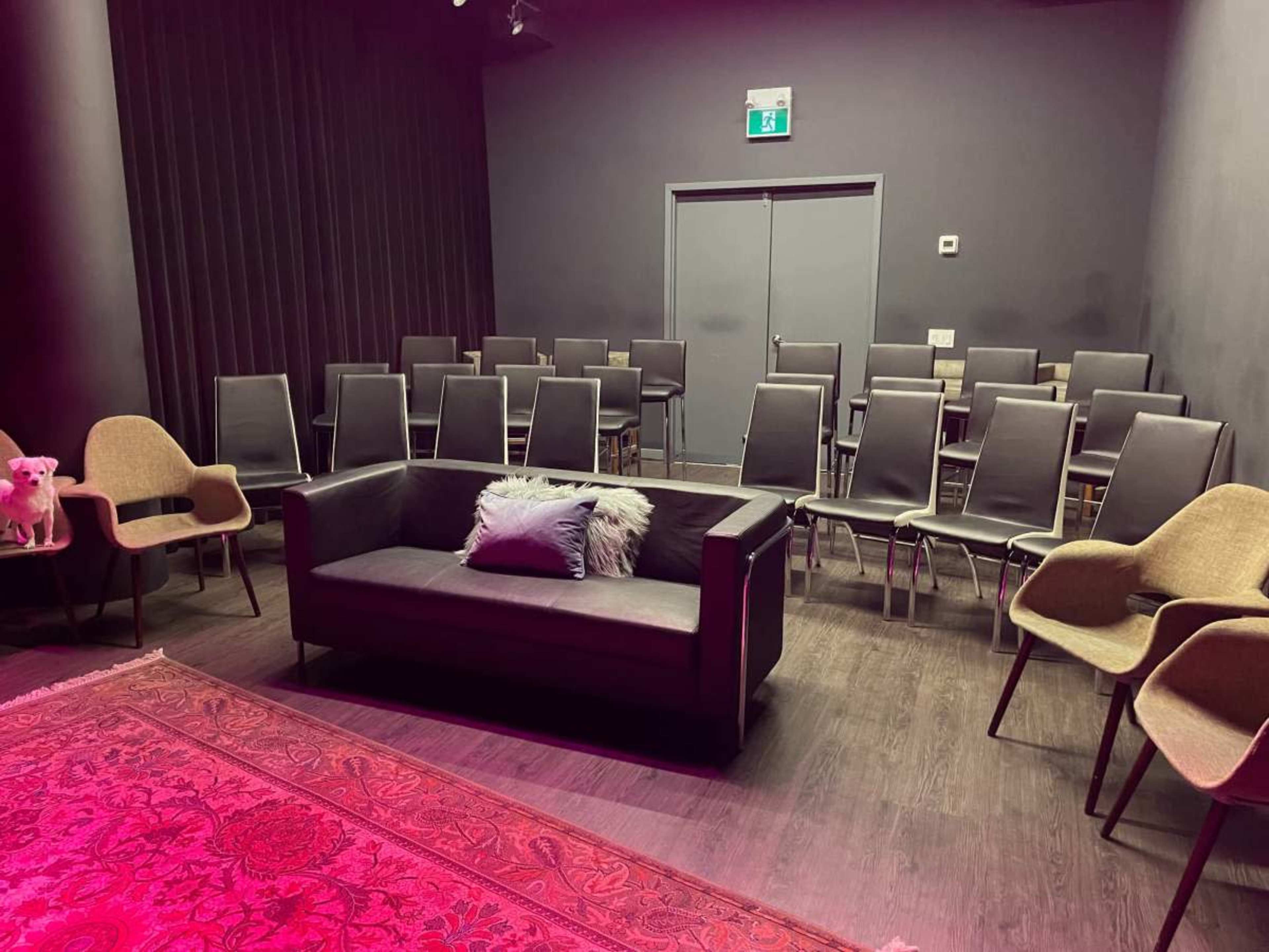 The image shows a small, empty performance space featuring a couch with a pillow, a patterned rug, and several rows of metal chairs.