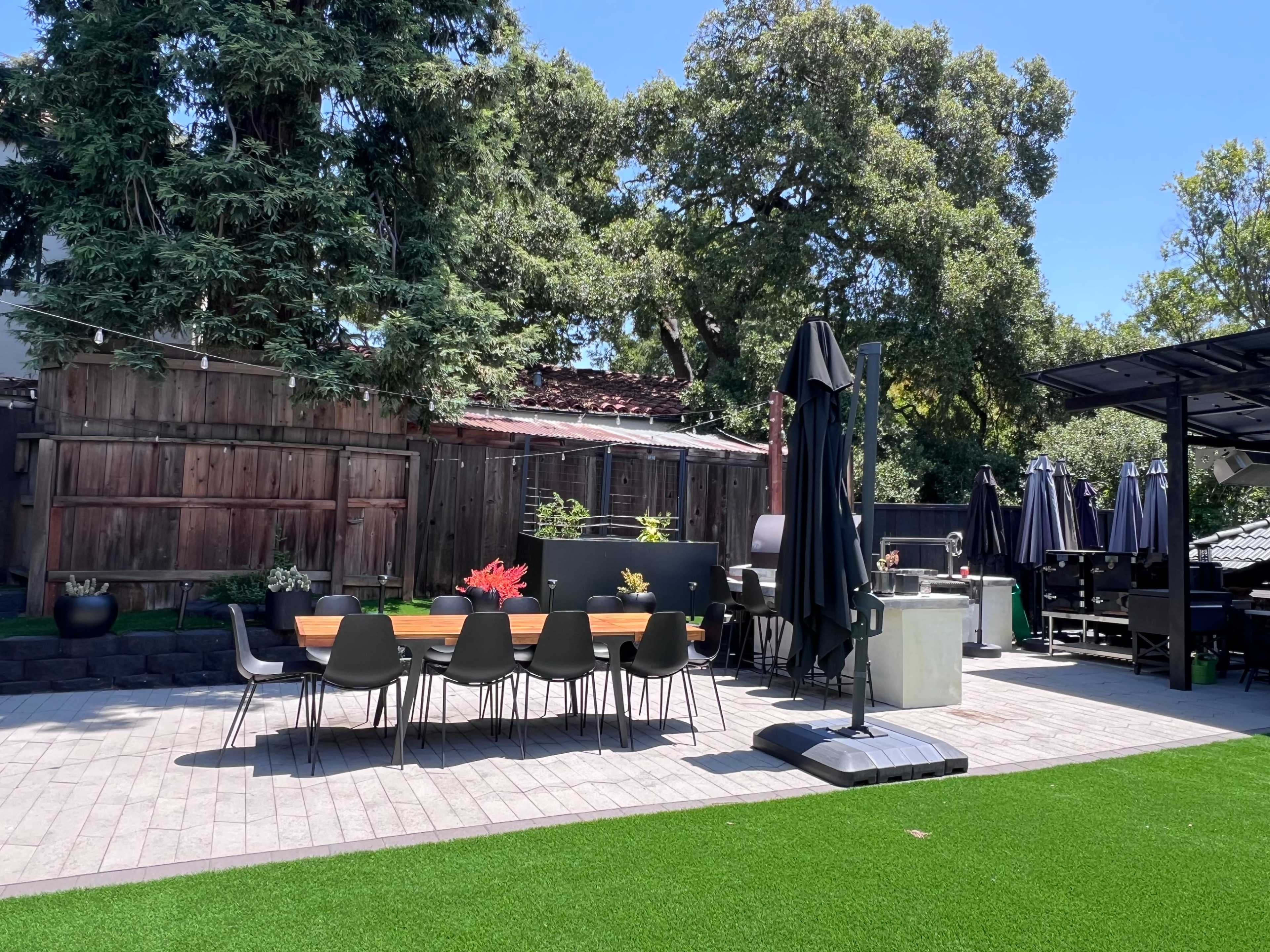 The image shows a landscaped outdoor patio with a long dining table, chairs, and several large umbrellas for shade, surrounded by greenery and trees.