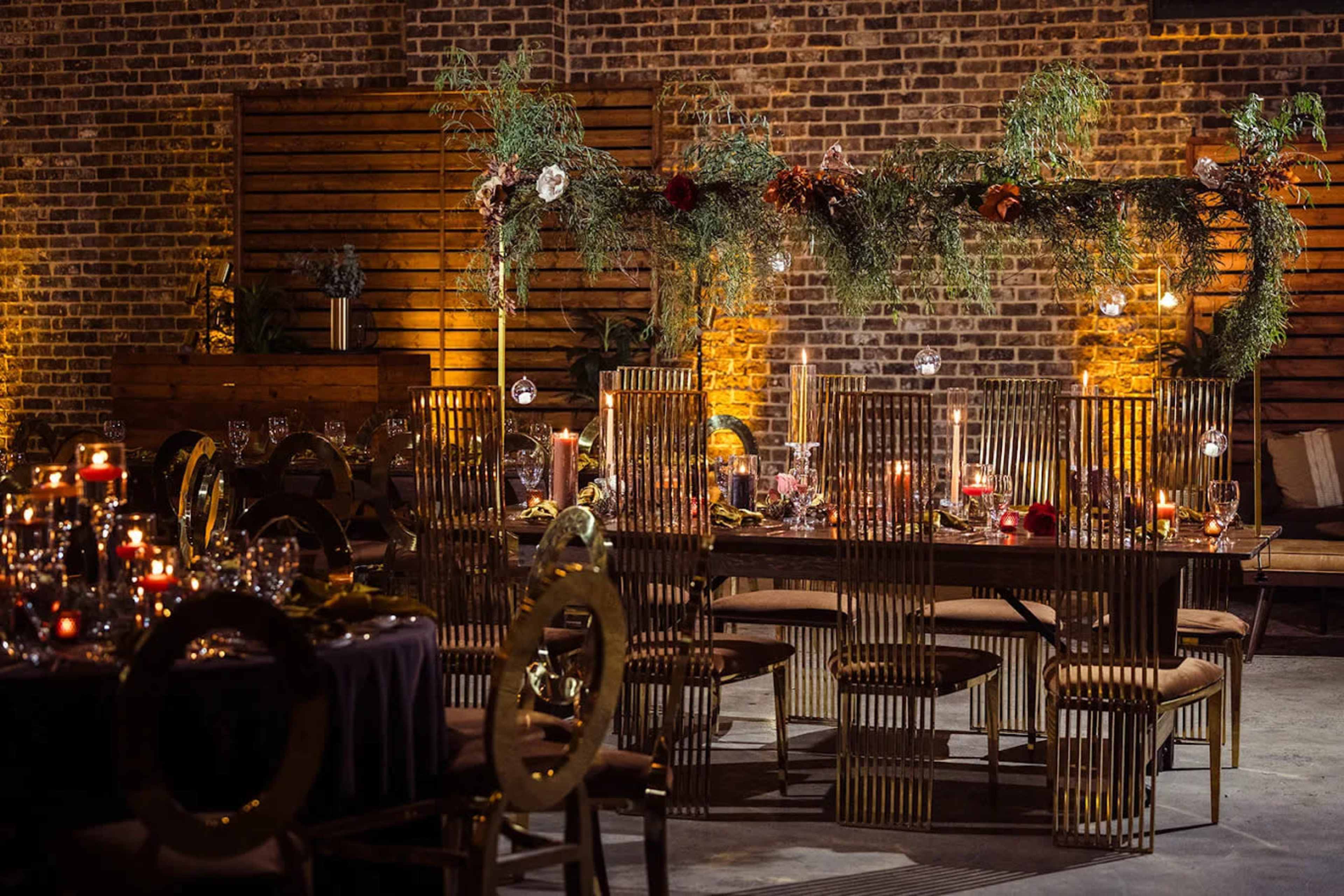 The image shows a decorated dining area with a long table set for an event, featuring candles, floral arrangements, and a brick wall backdrop.