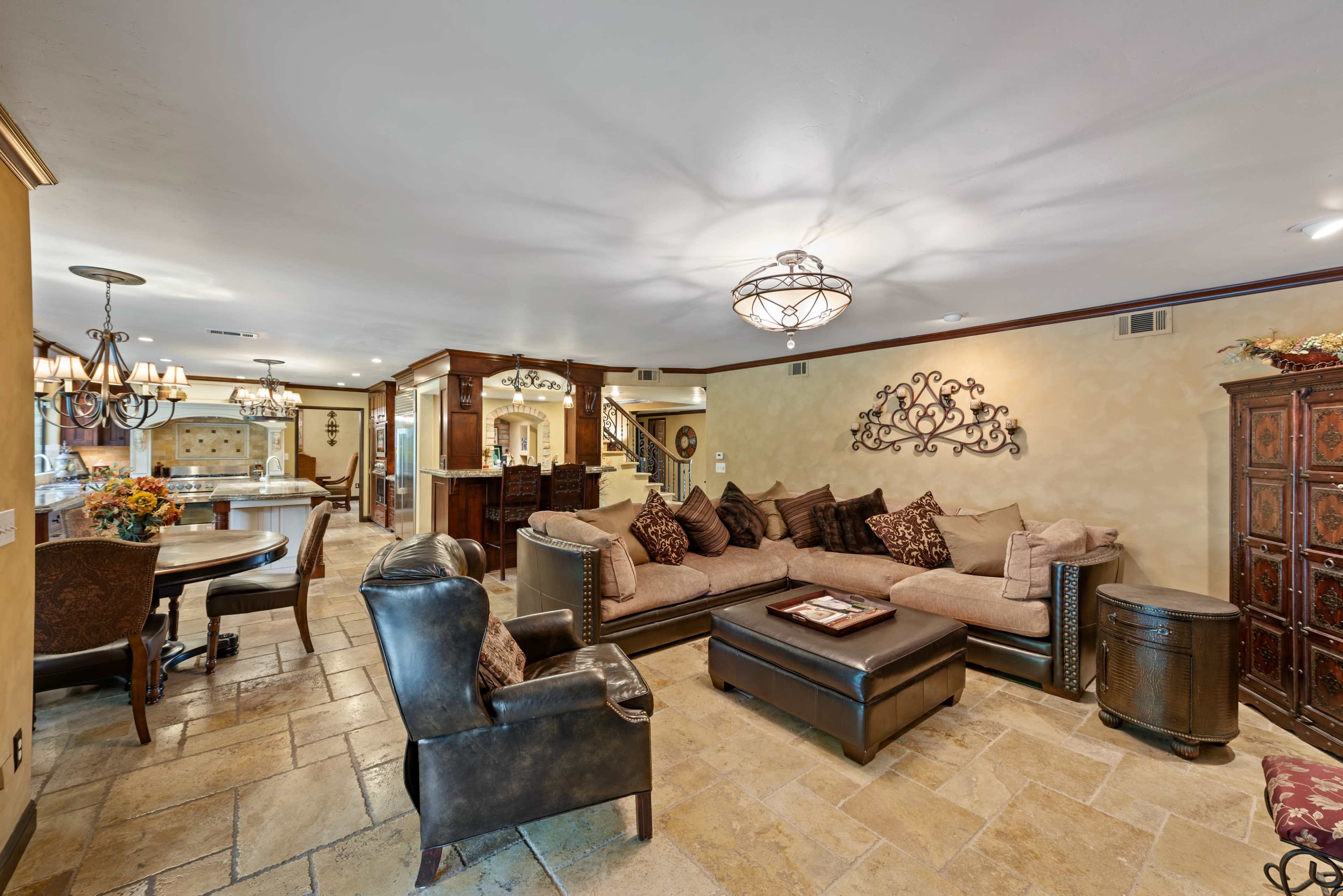 The image shows a spacious living room with a large sectional sofa, a coffee table, and decorative wall art, leading to a dining area and kitchen in the background.