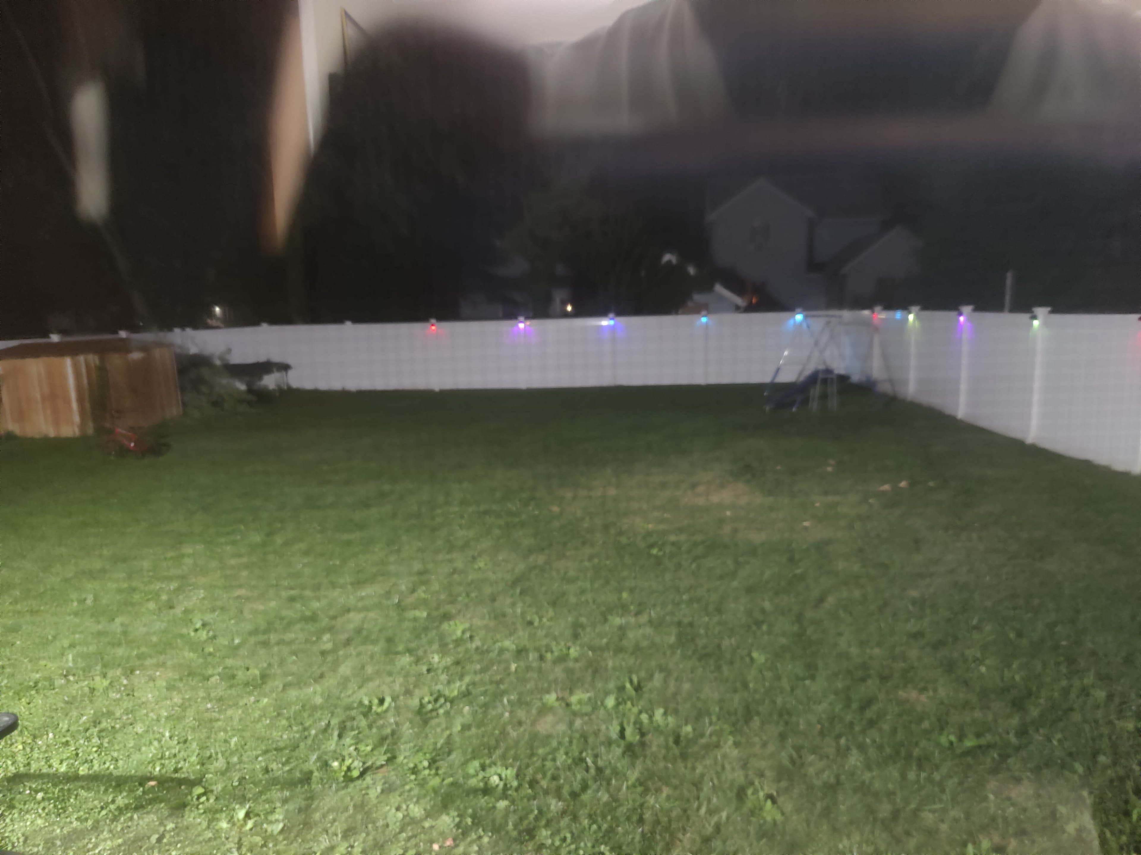 A backyard with a grassy area, a white fence, and colorful lights strung along the top.