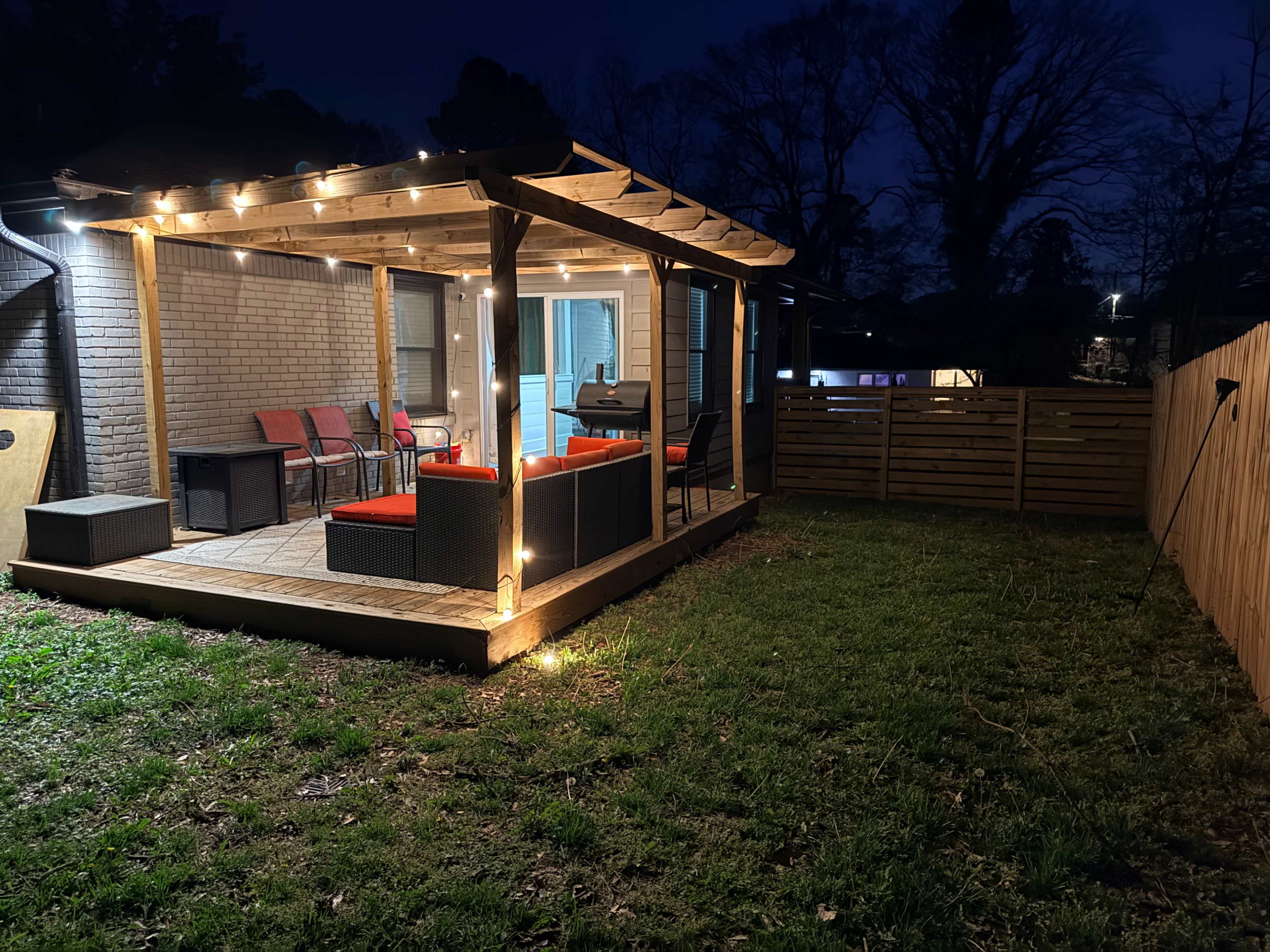 The image shows a well-lit outdoor patio with string lights, furnished with seating, and surrounded by grass and a wooden fence at night.