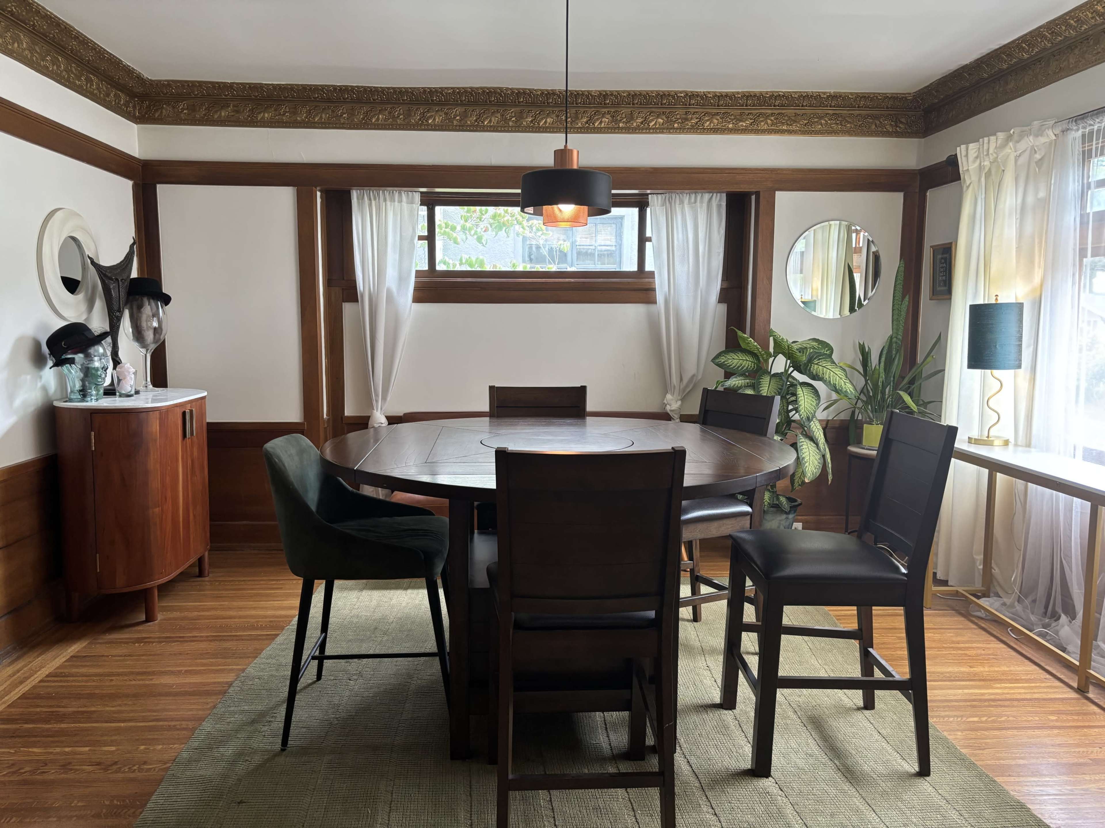 A round wooden dining table surrounded by tall chairs is set in a well-lit room with wooden paneling and large windows.