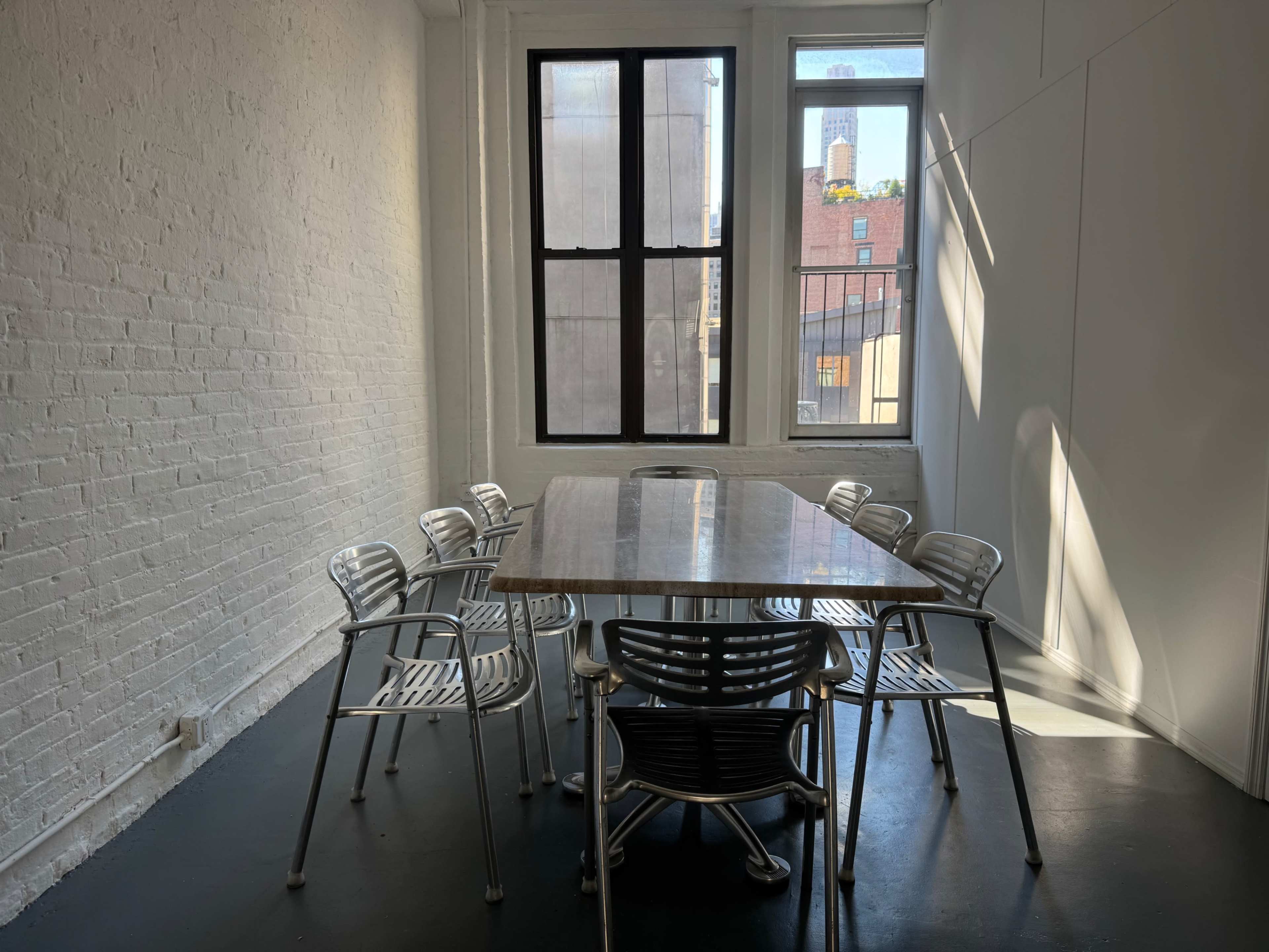 The image shows a modern conference room with a long table surrounded by metal chairs and large windows allowing natural light to enter.