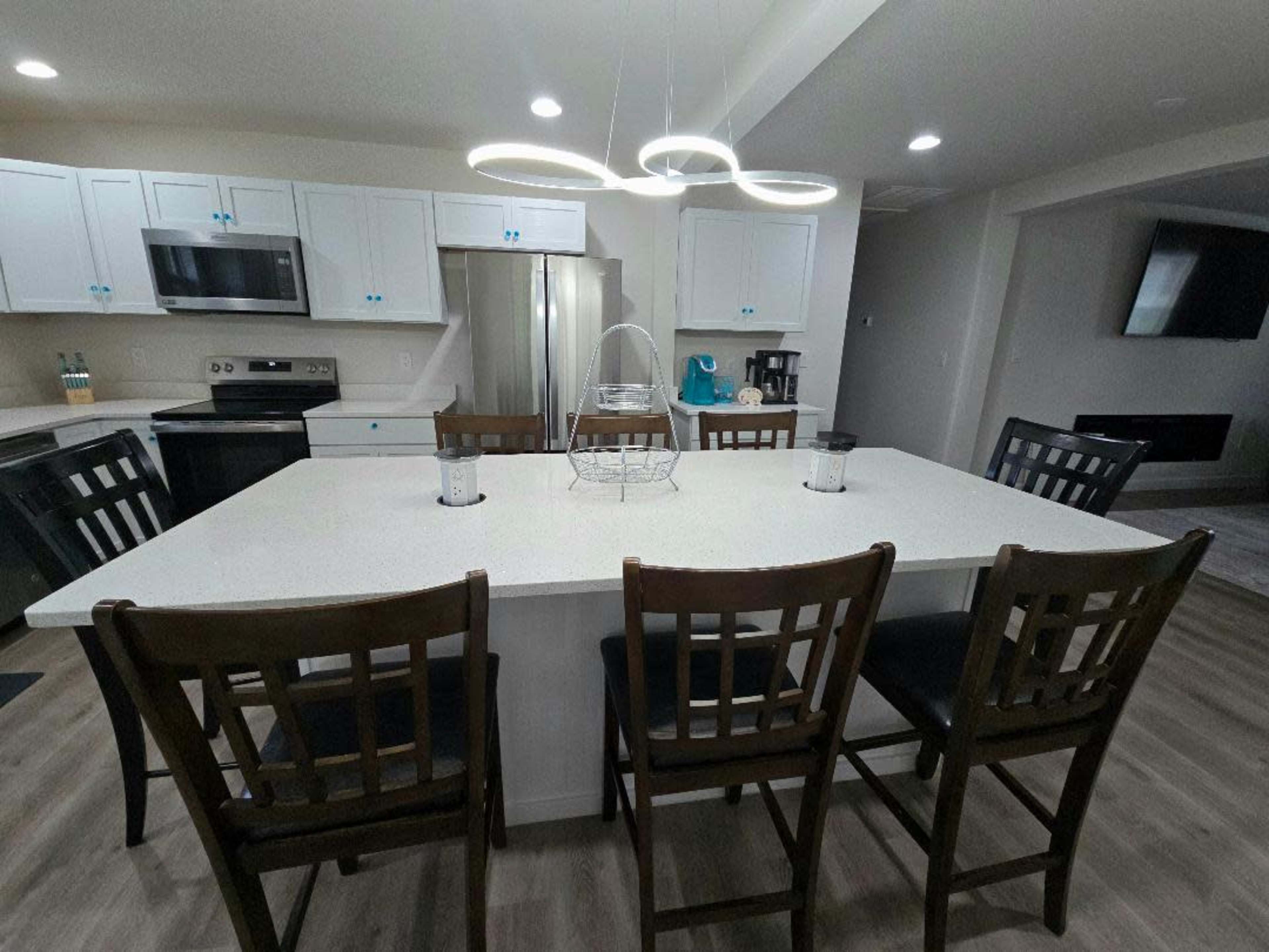 The image shows a modern kitchen with white cabinetry, a large island with seating for six, stainless steel appliances, and pendant lighting.