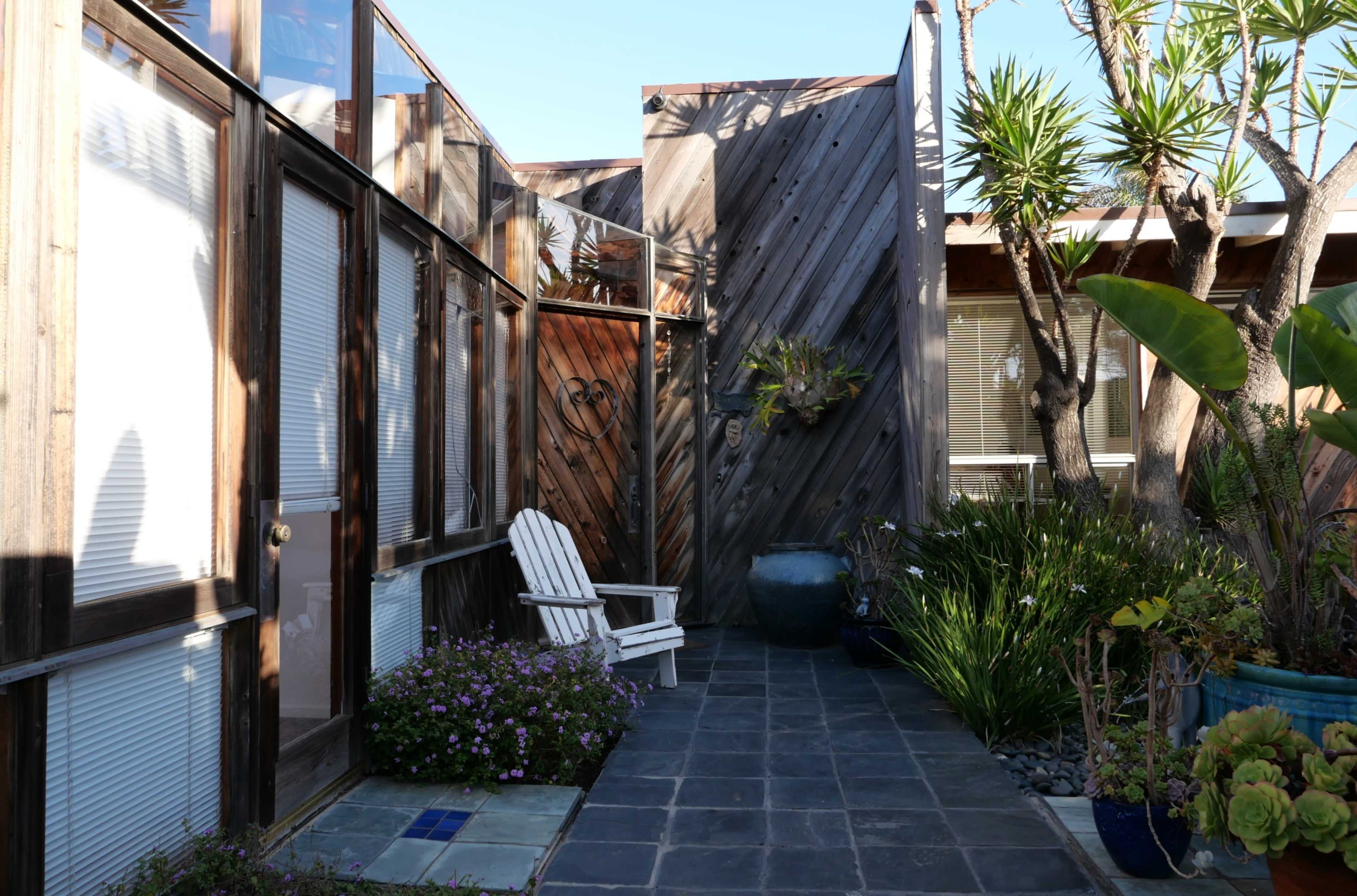 A pathway lined with plants and a blue pot, leading to a wooden structure with large glass windows and a white Adirondack chair.