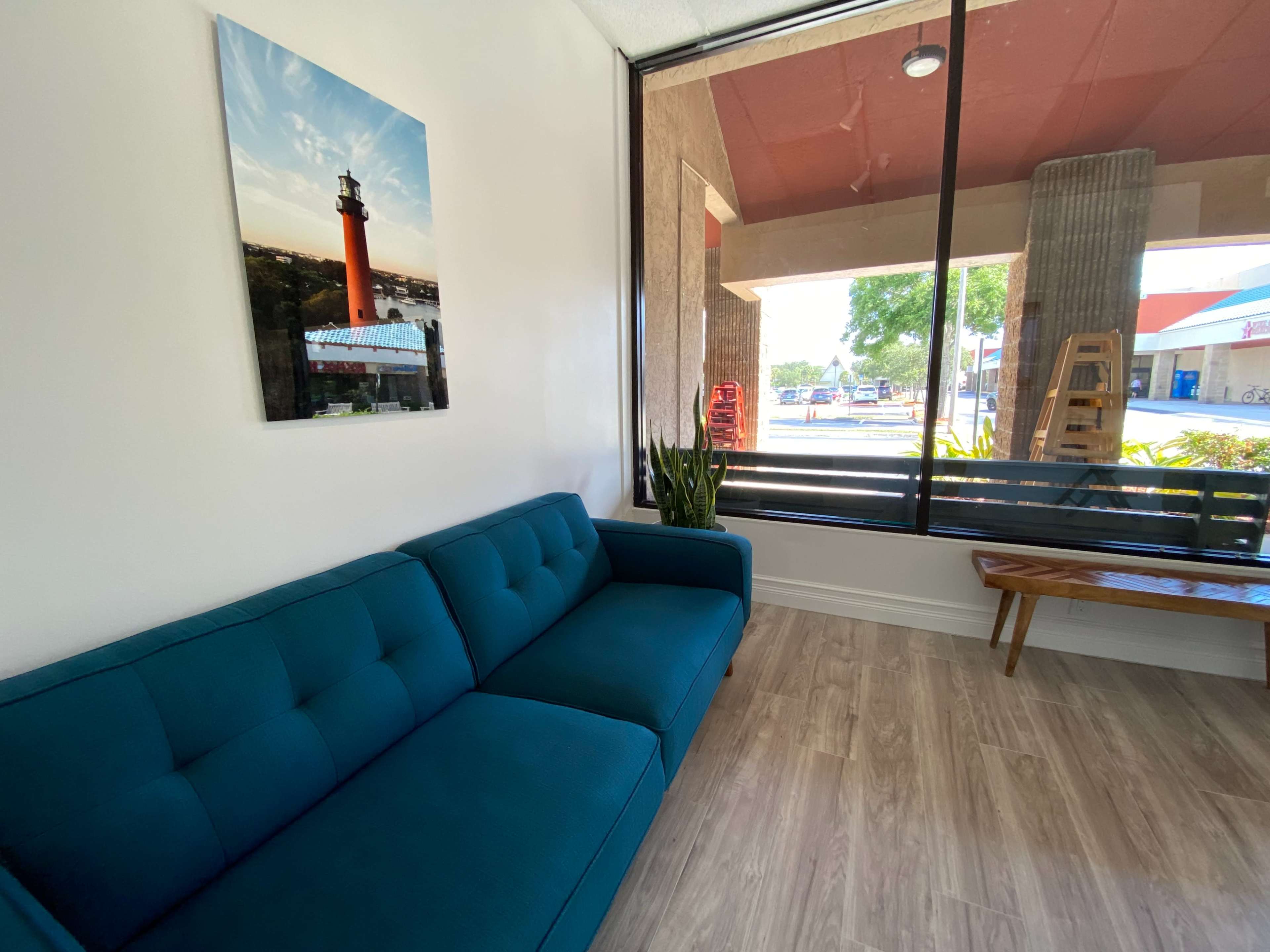 A waiting area with a teal couch, a wooden bench, and a large window overlooking a street, featuring a lighthouse picture on the wall.