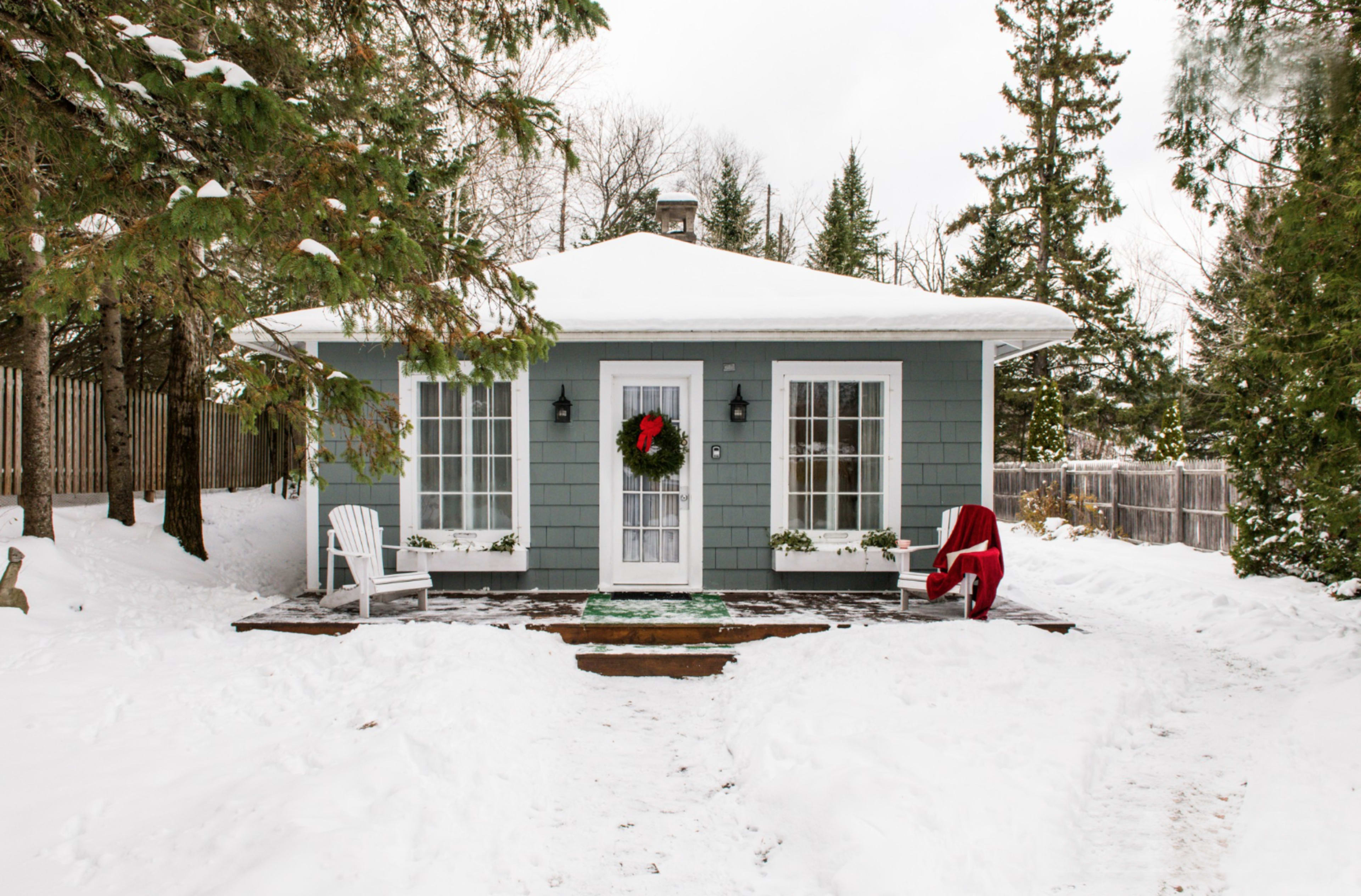 A small, snow-covered cottage with a green exterior features a festive wreath on the door and a red chair on the porch.