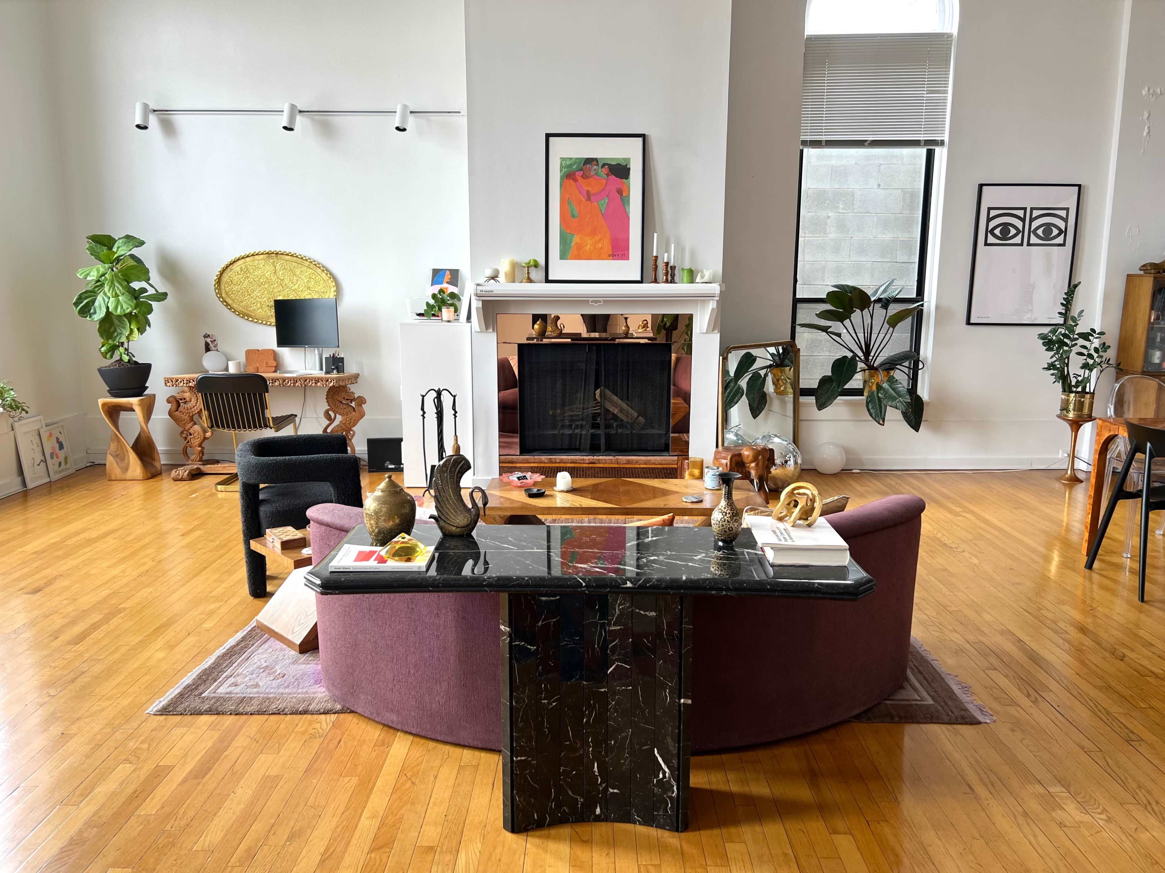 The image depicts a modern living room featuring a curved purple sofa, a black marble coffee table, various indoor plants, and a fireplace surrounded by artwork and decorative items.