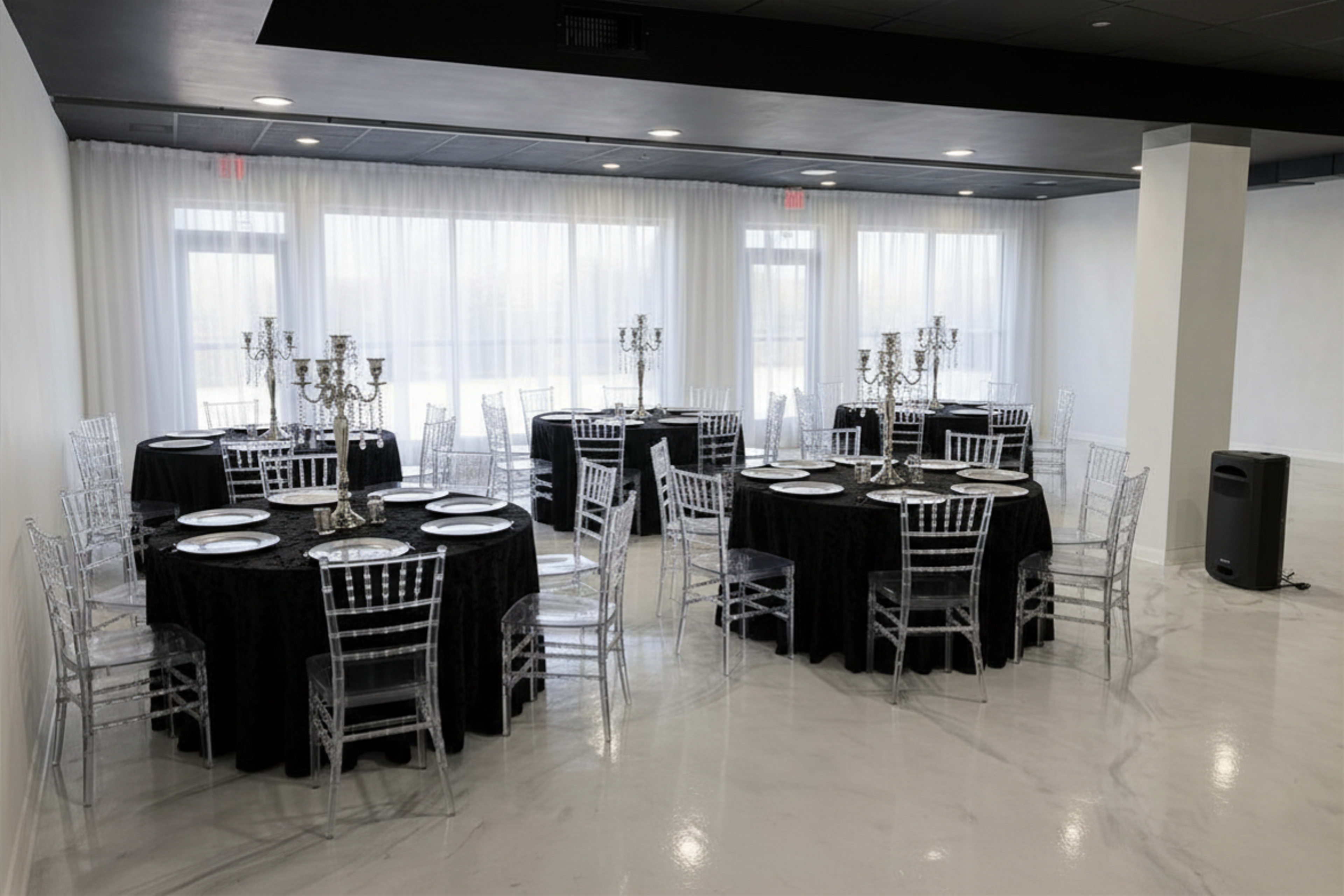 The image shows a spacious event room arranged with several round tables featuring black tablecloths, clear Chiavari chairs, and candelabras as centerpieces.