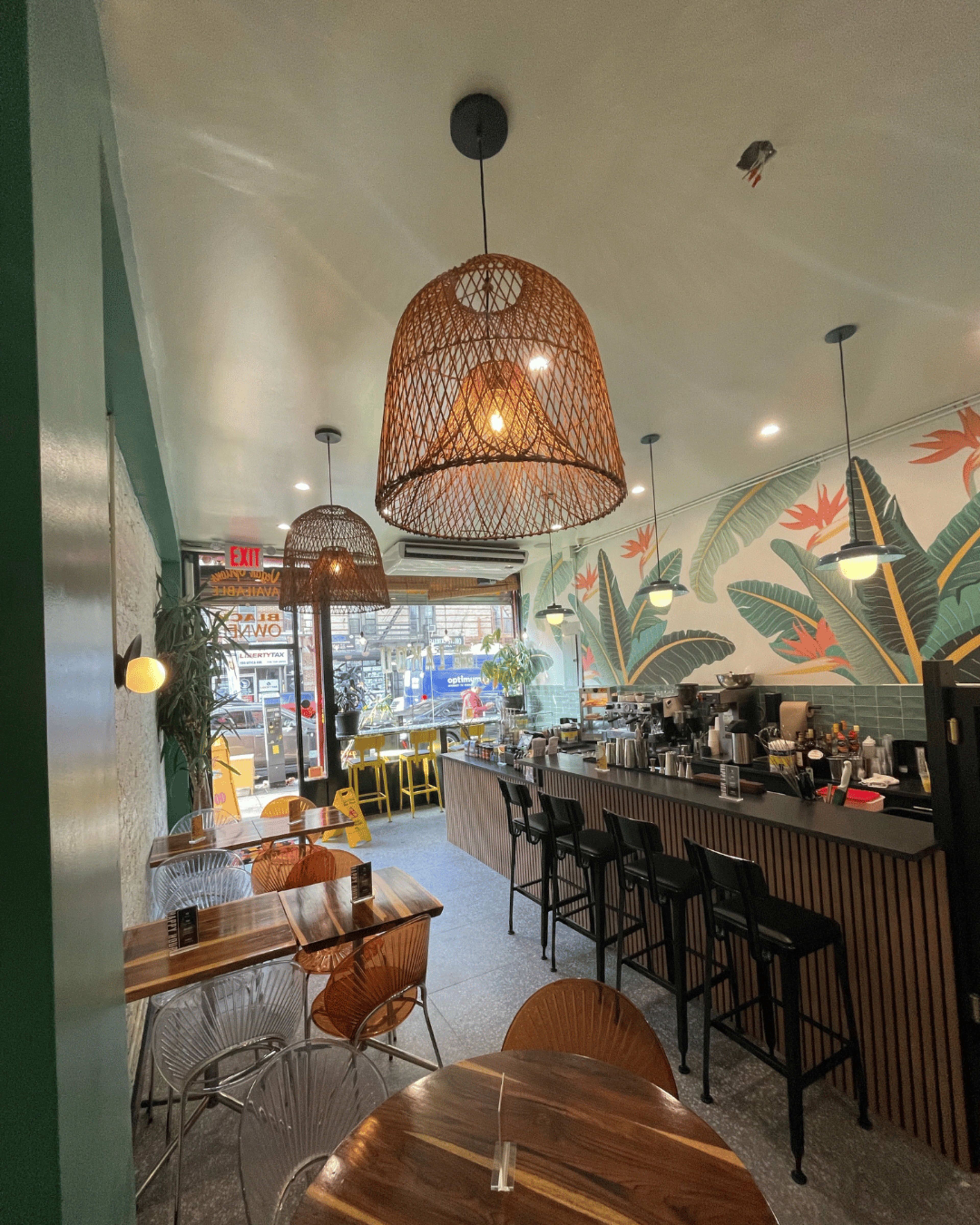 The image shows a cafe interior with a wooden counter, several tables, and decorative plants on the walls, illuminated by woven pendant lights.