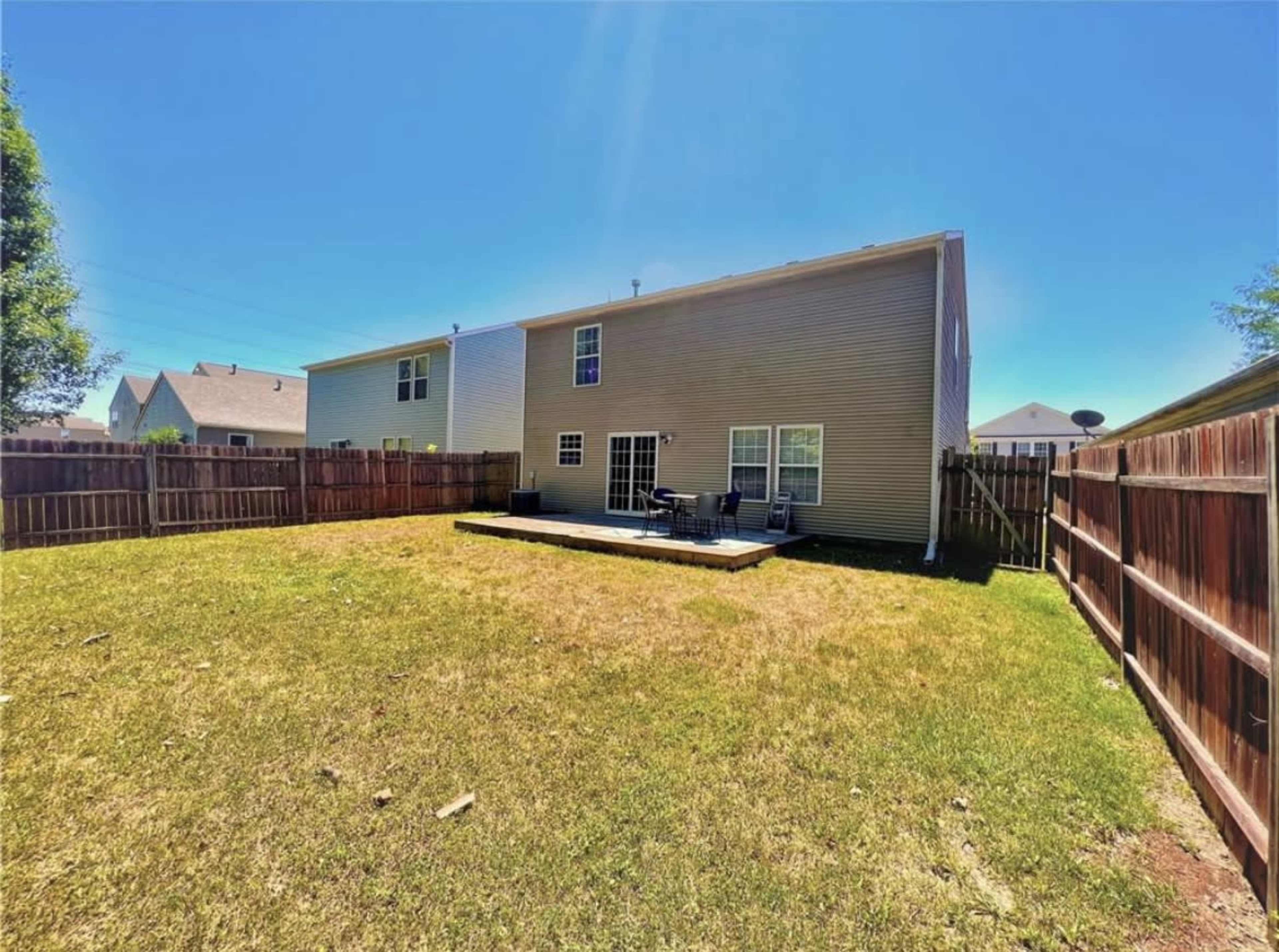 A backyard enclosed by a wooden fence, featuring a patch of grass and a small patio area with chairs.