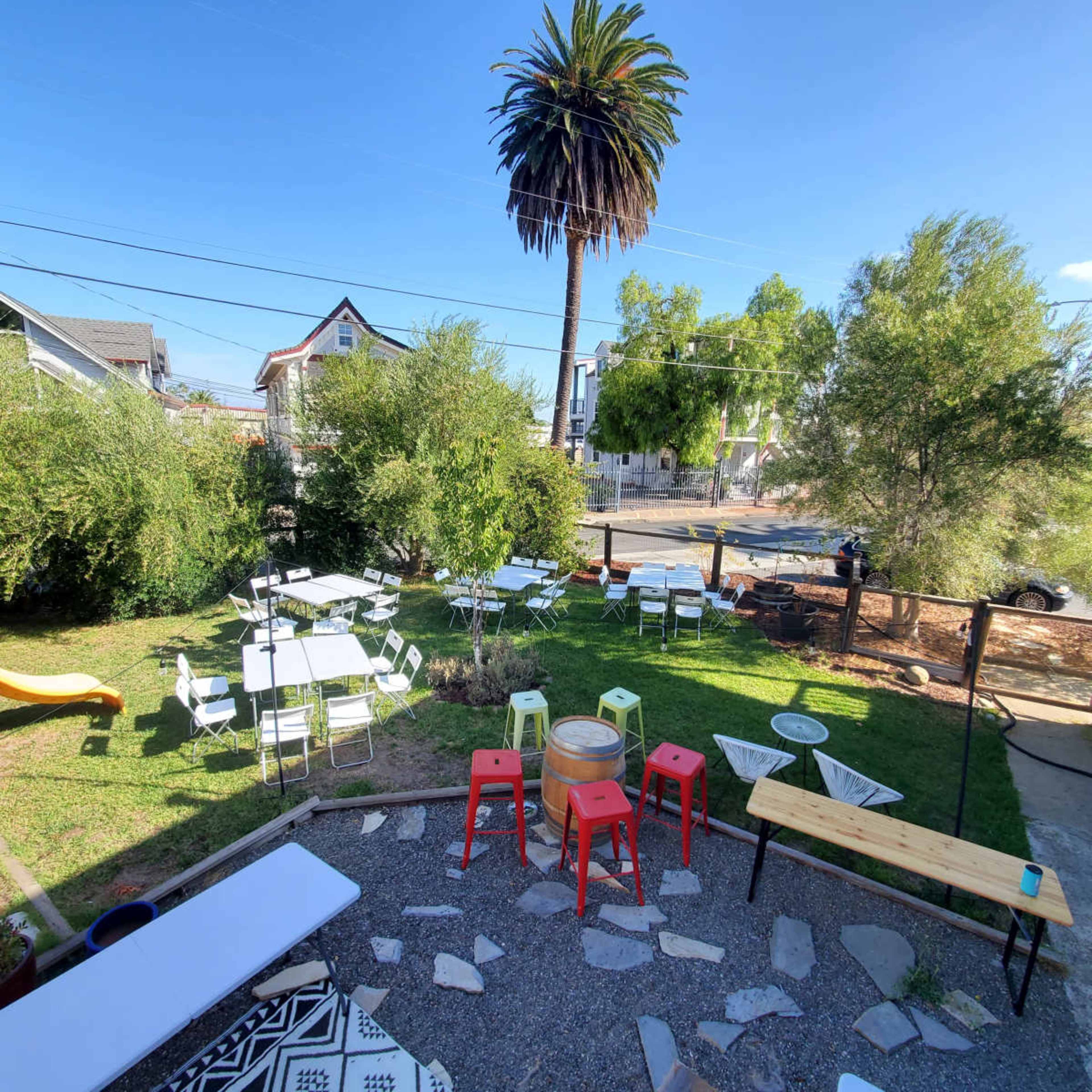 The image shows an outdoor seating area with several tables and chairs on a grassy lawn, surrounded by trees and a palm tree, with a view of a street in the background.