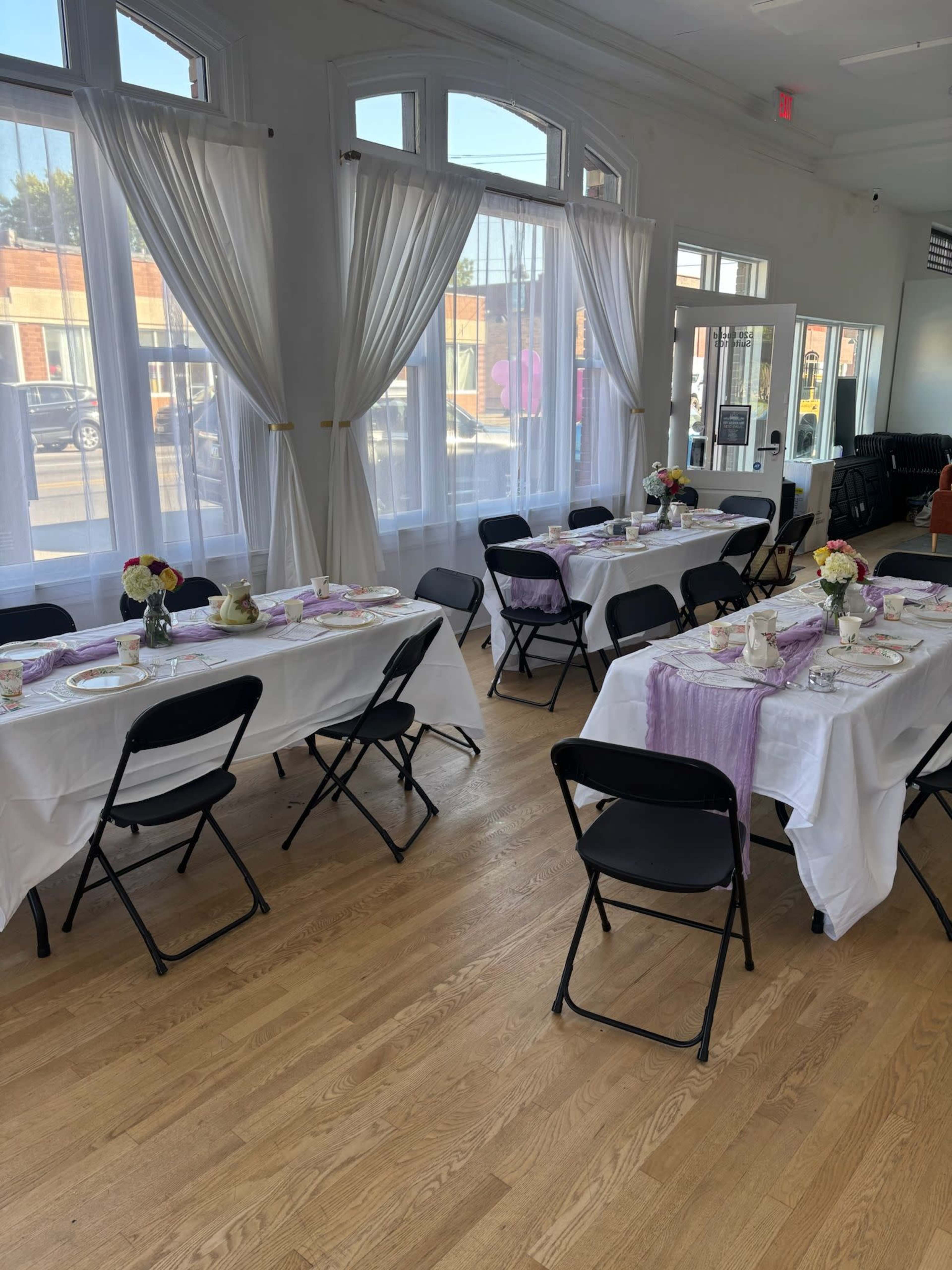 The image shows a venue with two sets of tables covered in white tablecloths, arranged for a dining event, with decorated centerpieces and natural light streaming through the windows.