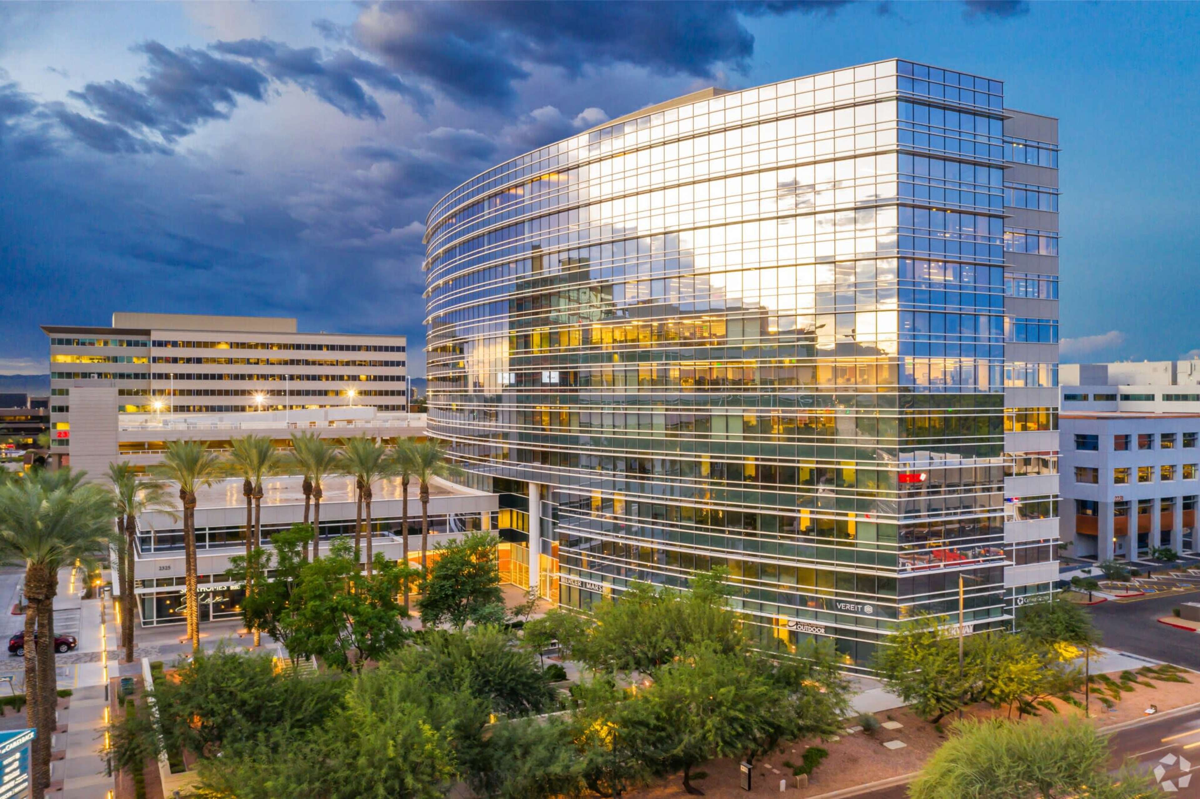 A modern glass office building surrounded by palm trees and other structures under a cloudy sky at dusk.