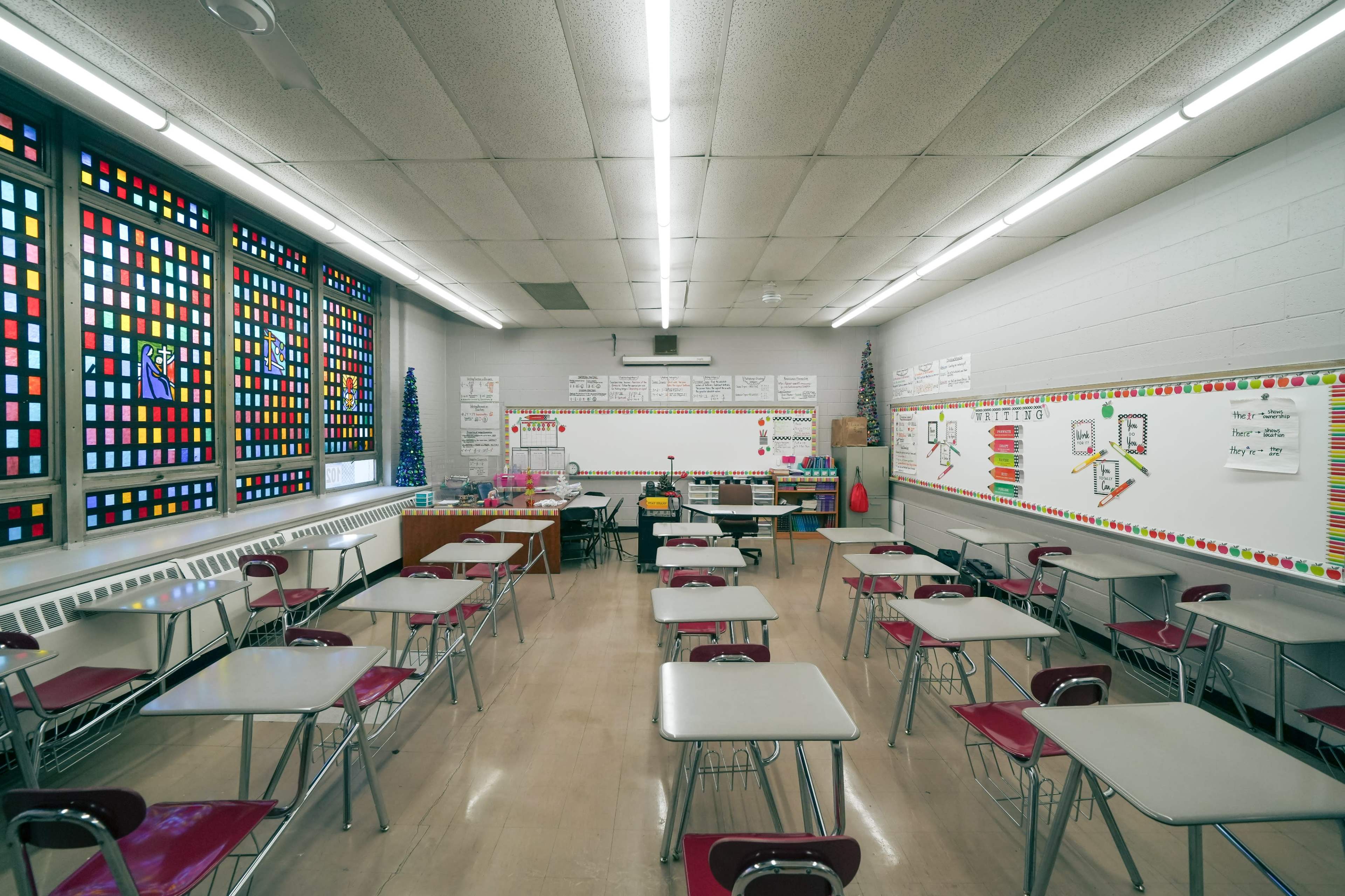 A classroom is arranged with desks facing a whiteboard, featuring colorful stained glass windows and educational materials on the walls.