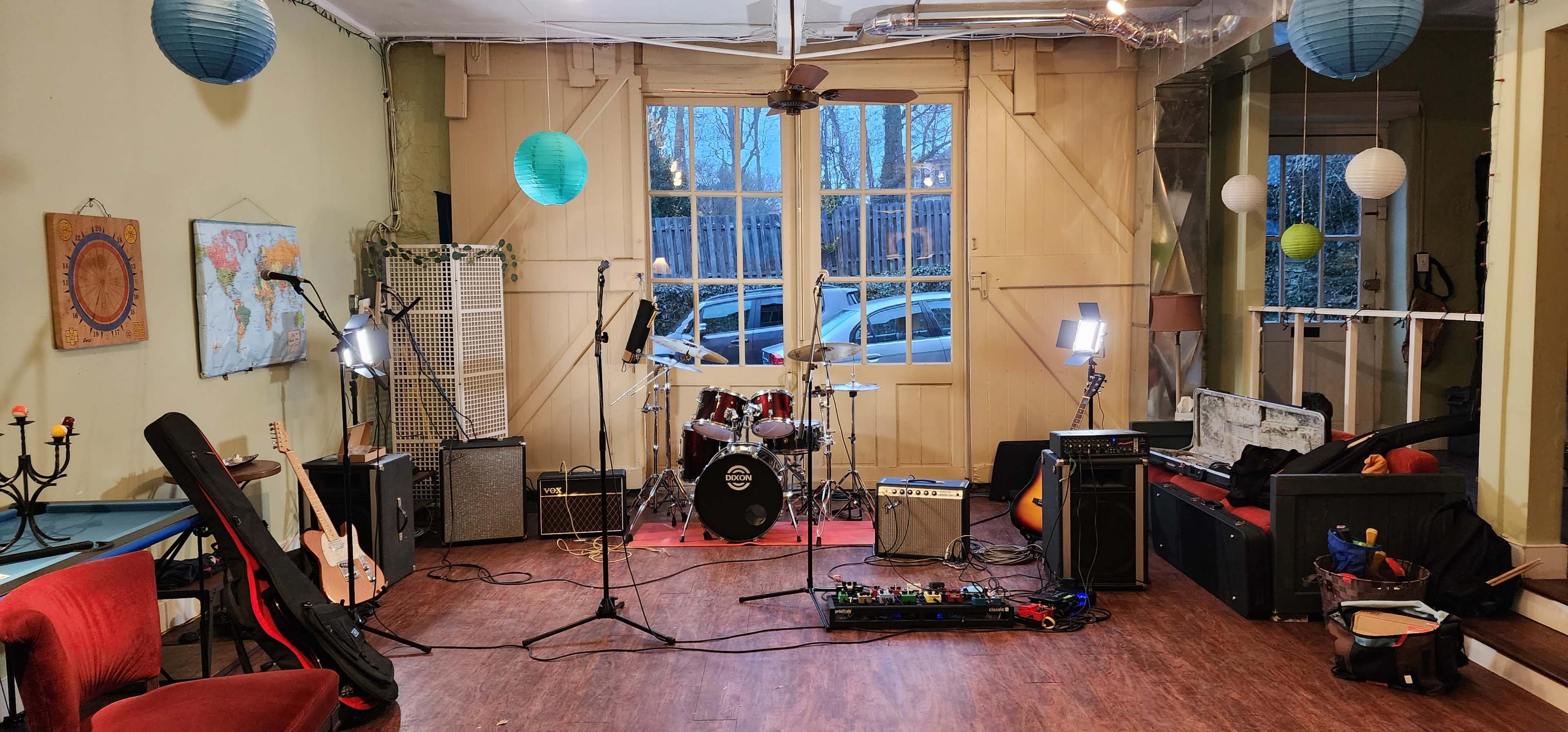 The image shows a music rehearsal space with a drum set, amplifiers, and various instruments arranged around a stage area near large windows.