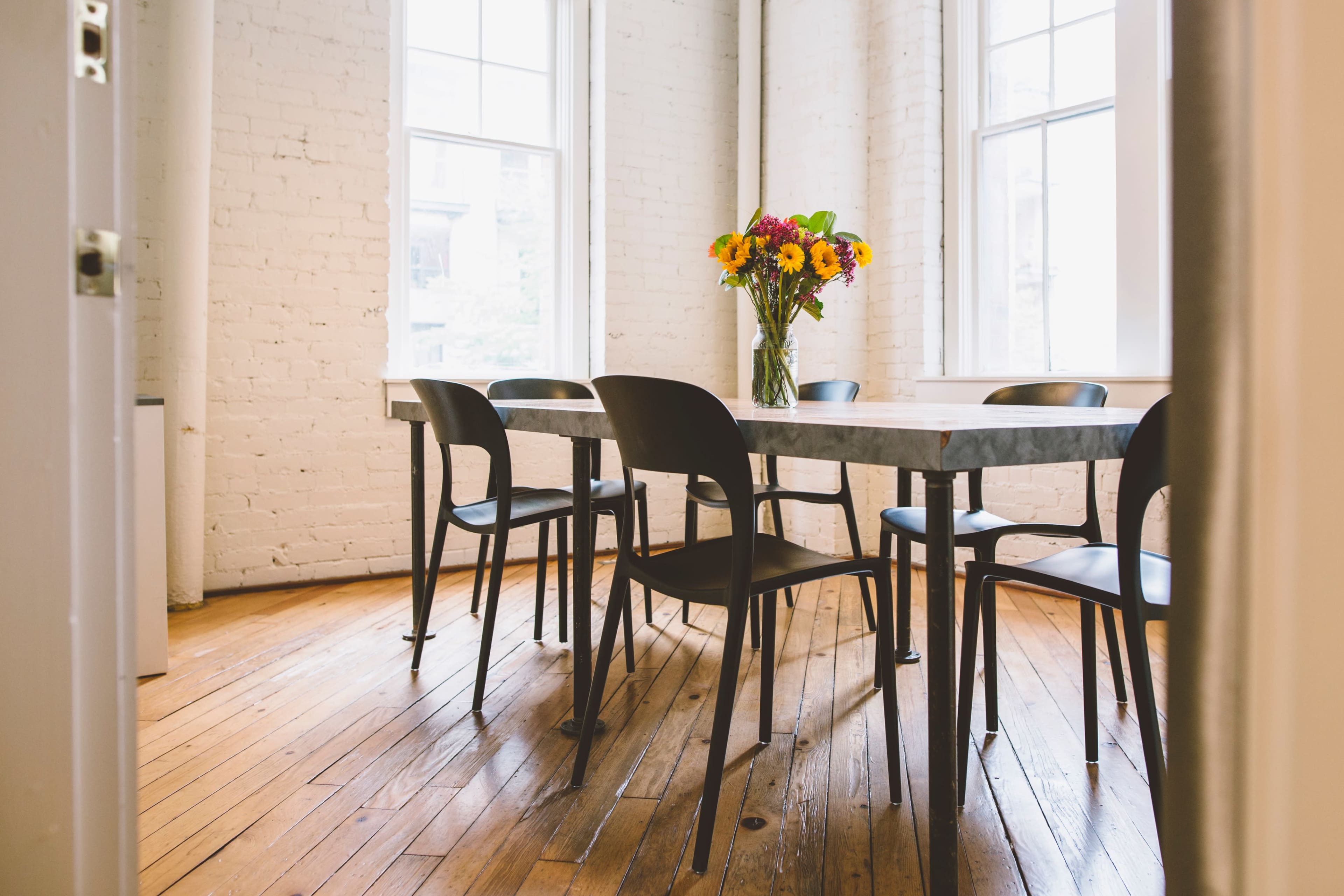 A table with six black chairs and a vase of flowers is placed in a room with white brick walls and large windows.
