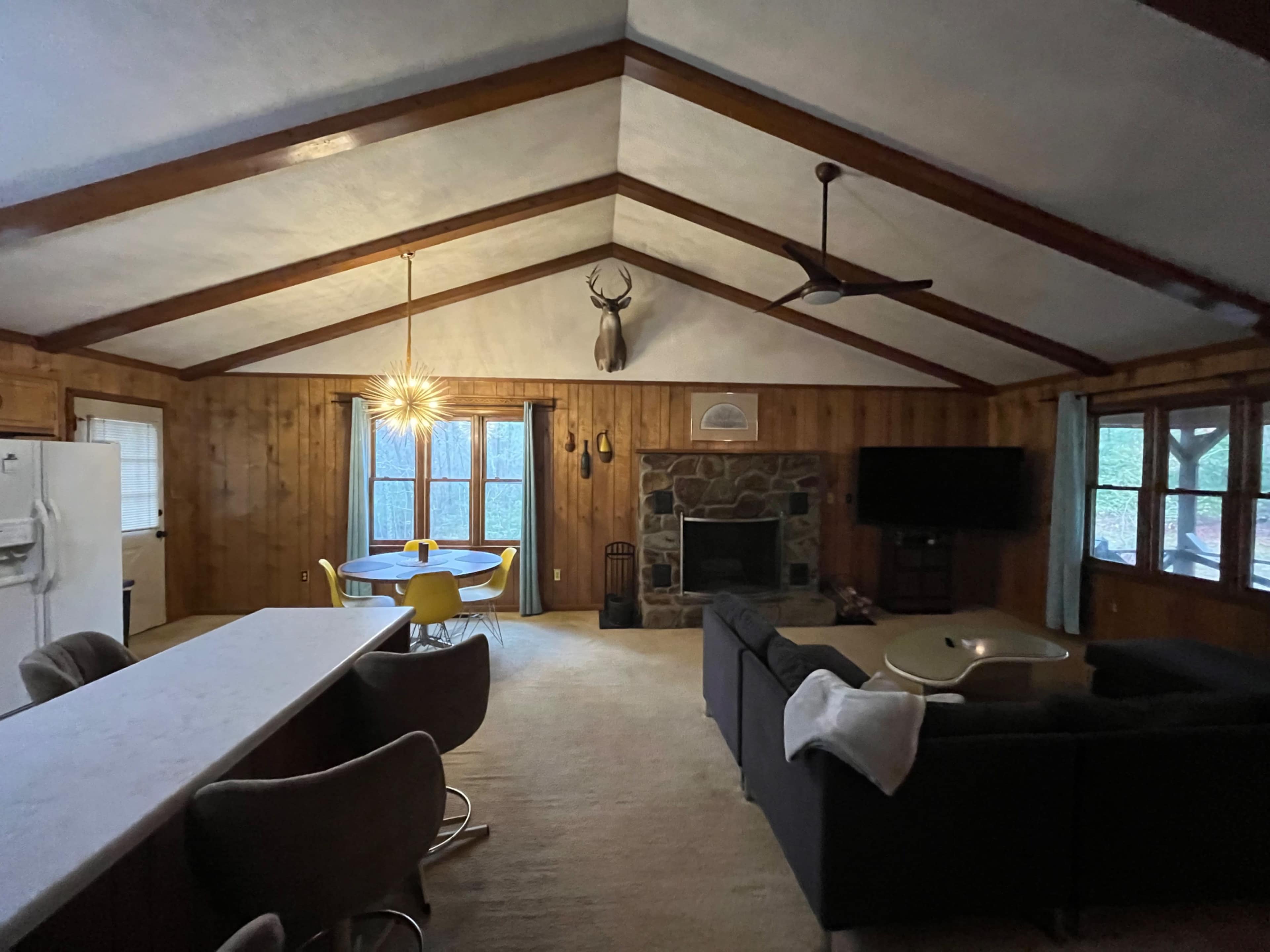 A spacious living area featuring a stone fireplace, large windows, a kitchen bar, and a dining table with yellow chairs.