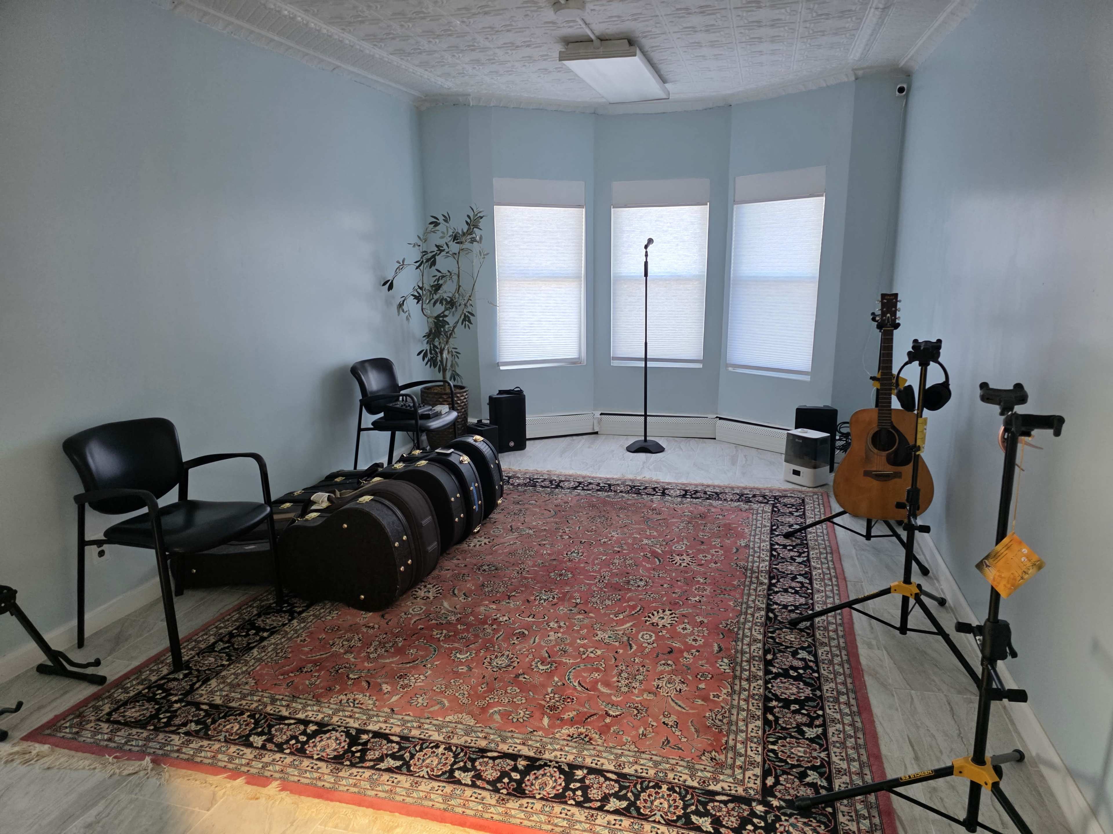 The room features a light blue wall, a patterned area rug, a microphone stand in the corner, two black chairs, several musical instruments, and large windows with natural light.
