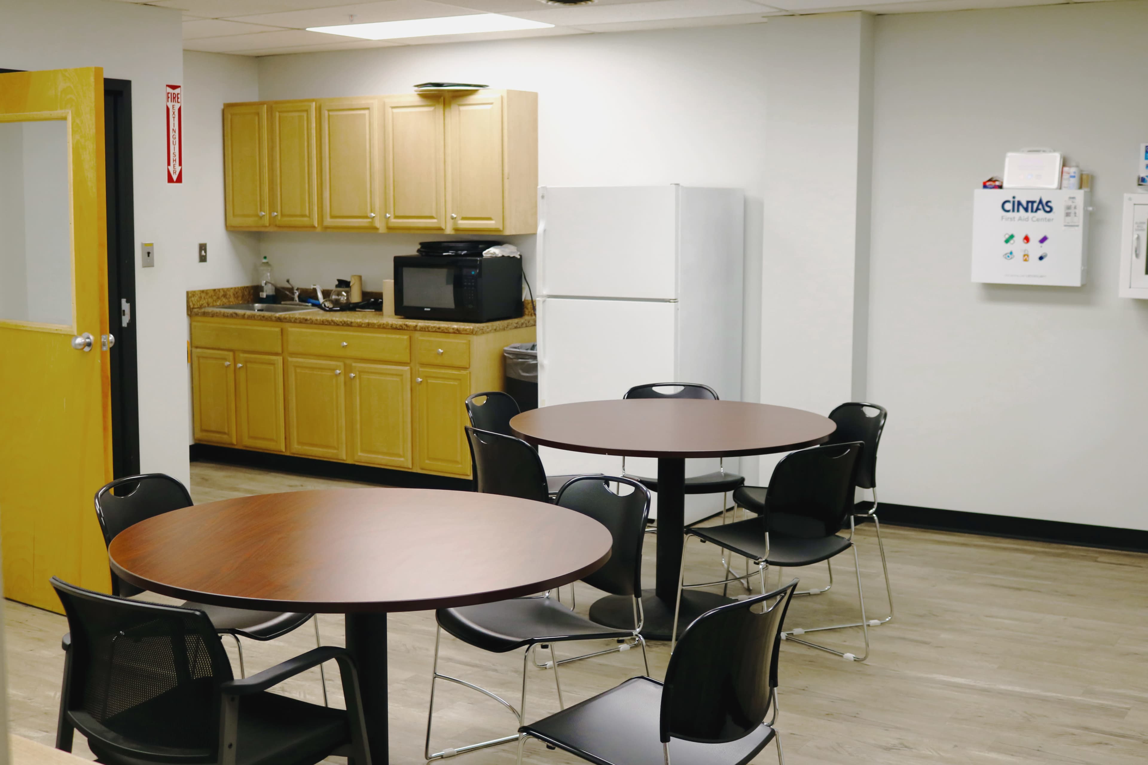 A break room with a kitchenette featuring cabinets, a refrigerator, and a microwave, alongside two tables surrounded by chairs.