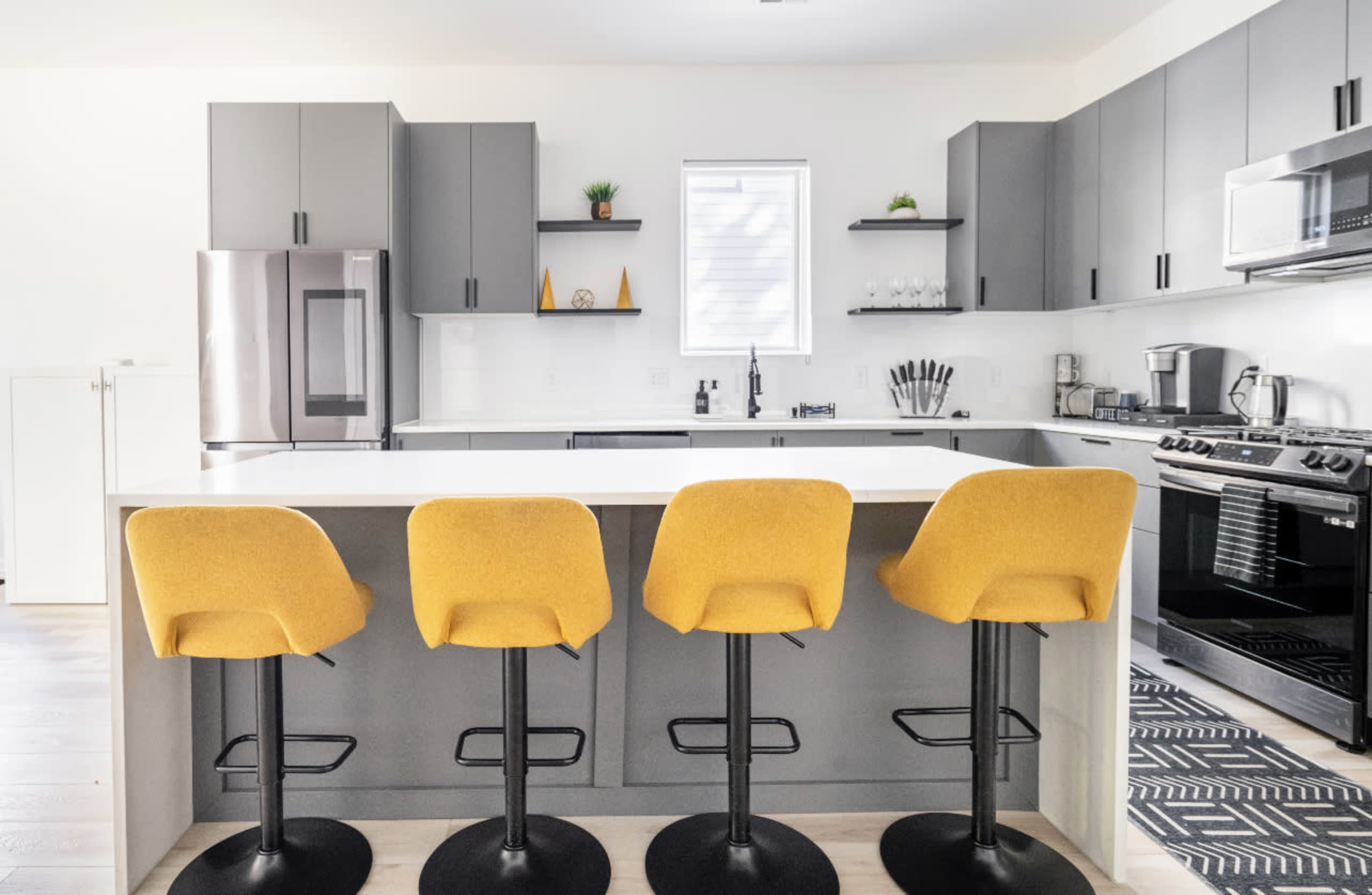 A modern kitchen features a central island with four yellow bar stools, sleek cabinetry, and stainless steel appliances.