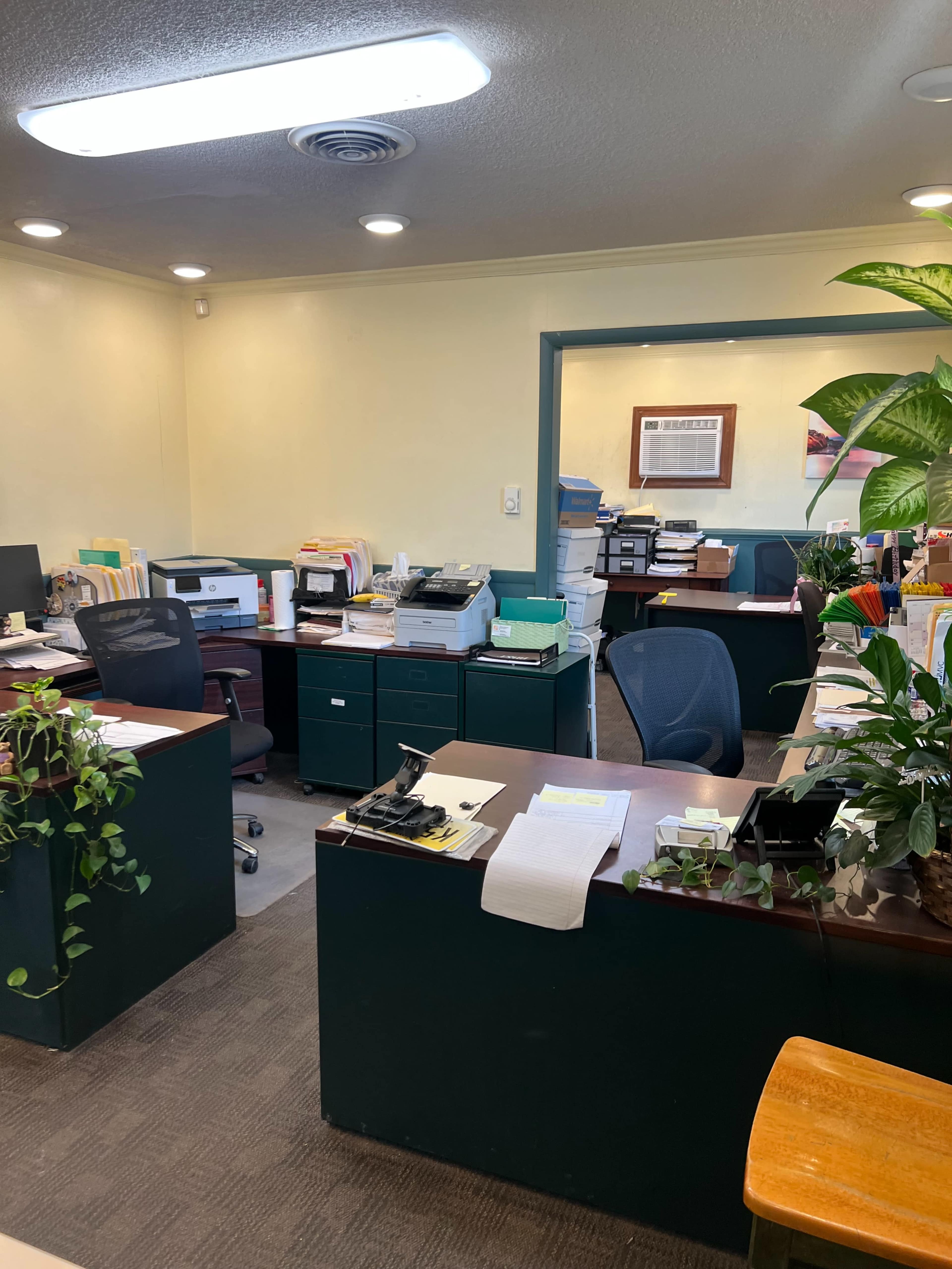 The image shows an office space with multiple desks, printers, filing cabinets, and plants arranged in a well-organized manner.