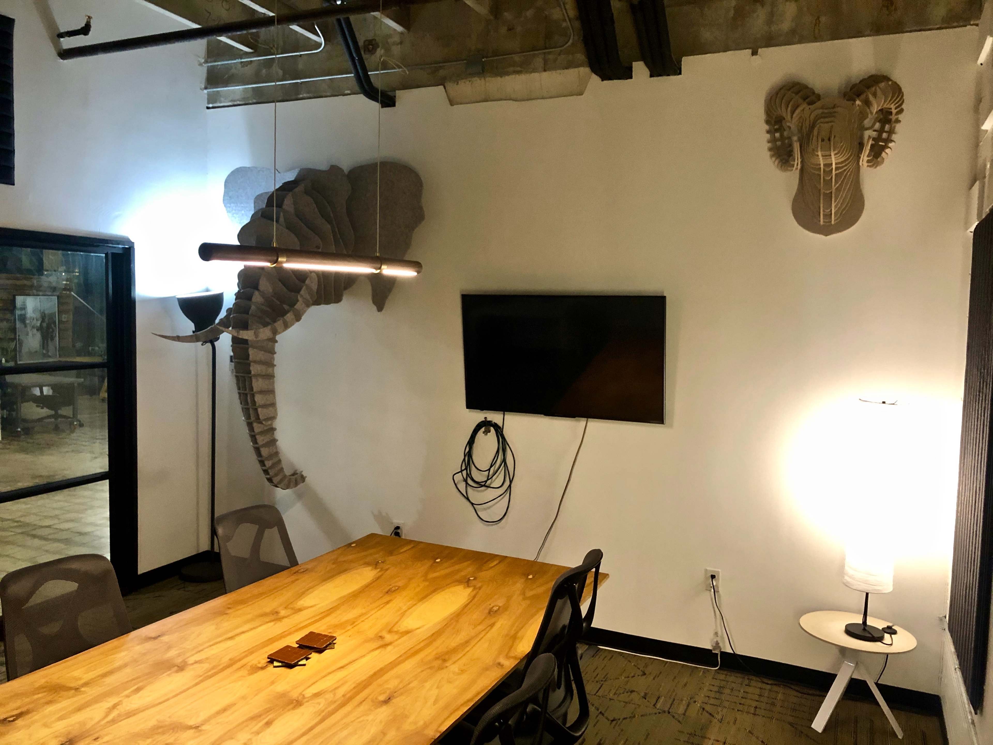 A modern conference room features a wooden table, an overhead light, a wall-mounted TV, and decorative elephant sculptures on the walls.